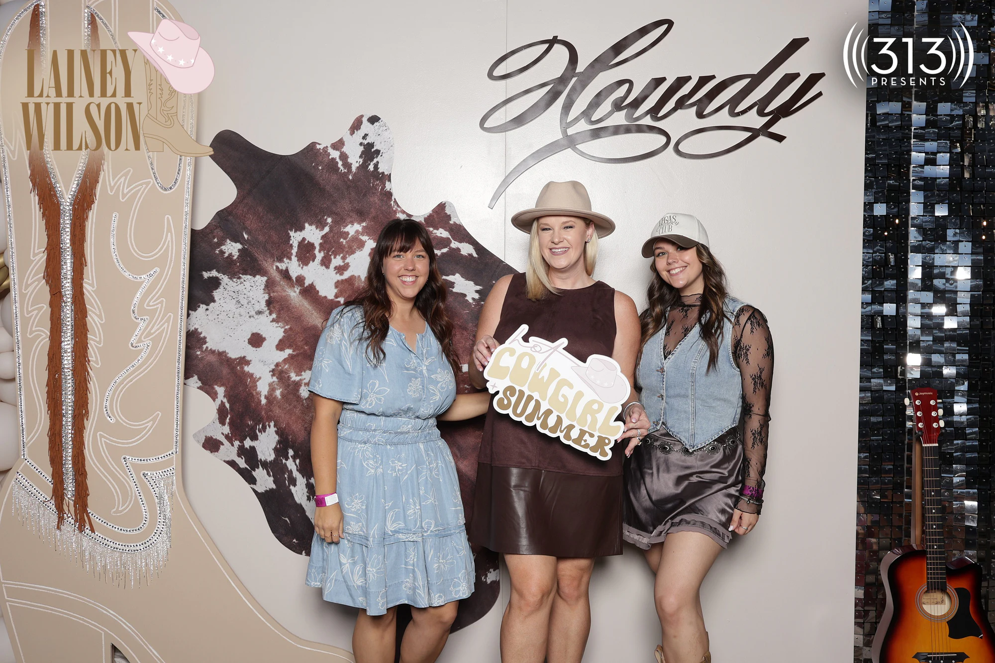 Three women holding the Cowgirl Summer sign against the cowhide backdrop at 313 Presents Concert Activations