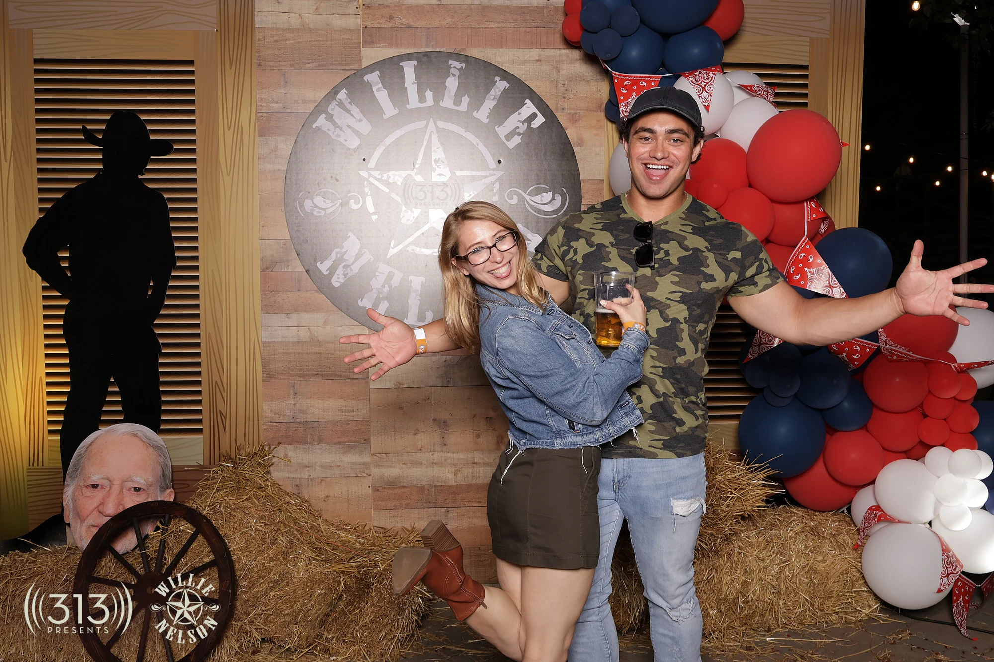 Couple mid-cheer, arms wide, the Willie Nelson sign centered behind them at 313 Presents Concert Activations