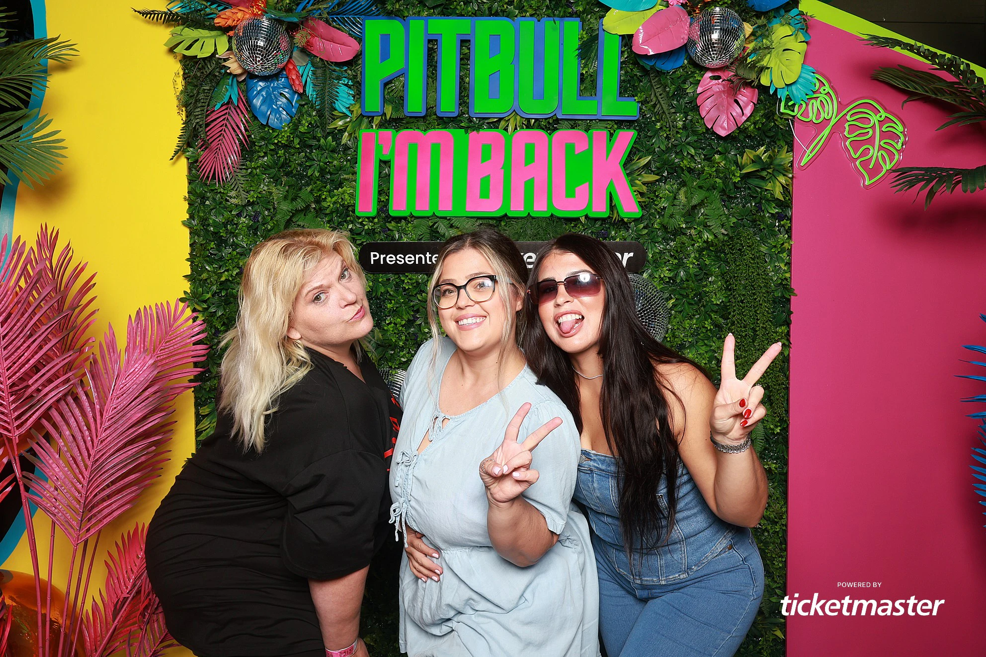 Three guests at the greenery wall, two of them throwing peace signs at Pitbull, Presented by Ticketmaster