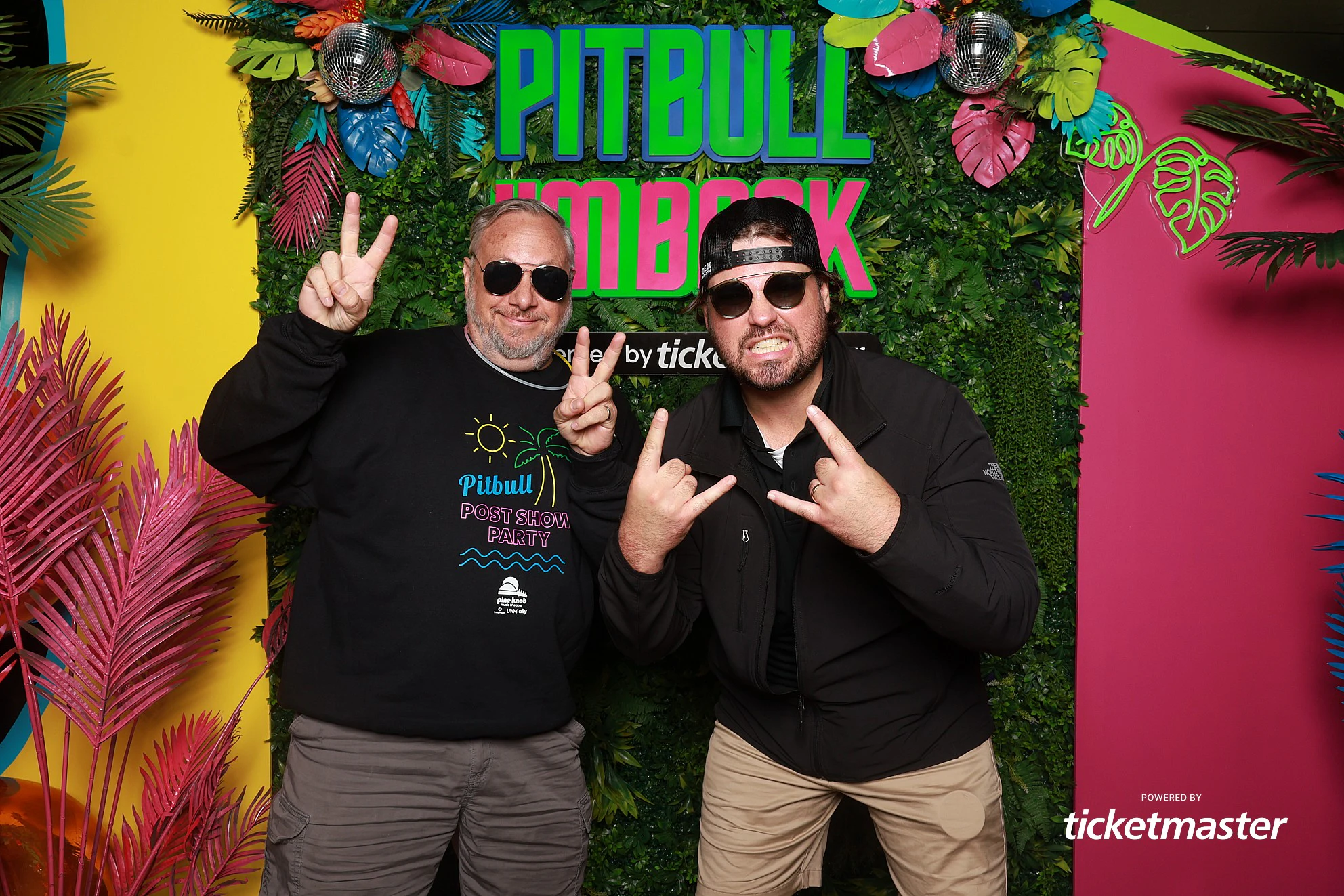 Two guests throwing peace signs and rock horns at the greenery wall at Pitbull, Presented by Ticketmaster