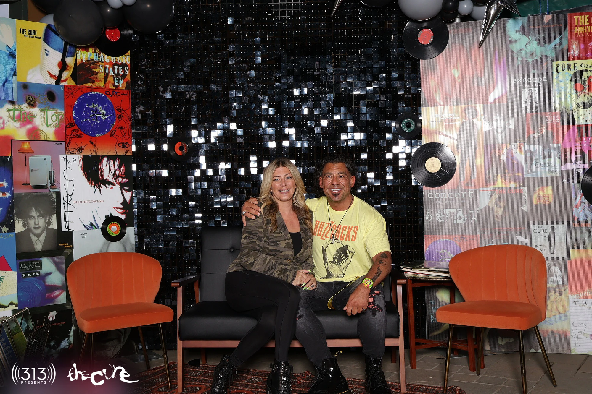 A couple on the couch in front of the sequin wall, album covers either side at The Cure VIP Party with 313 Presents