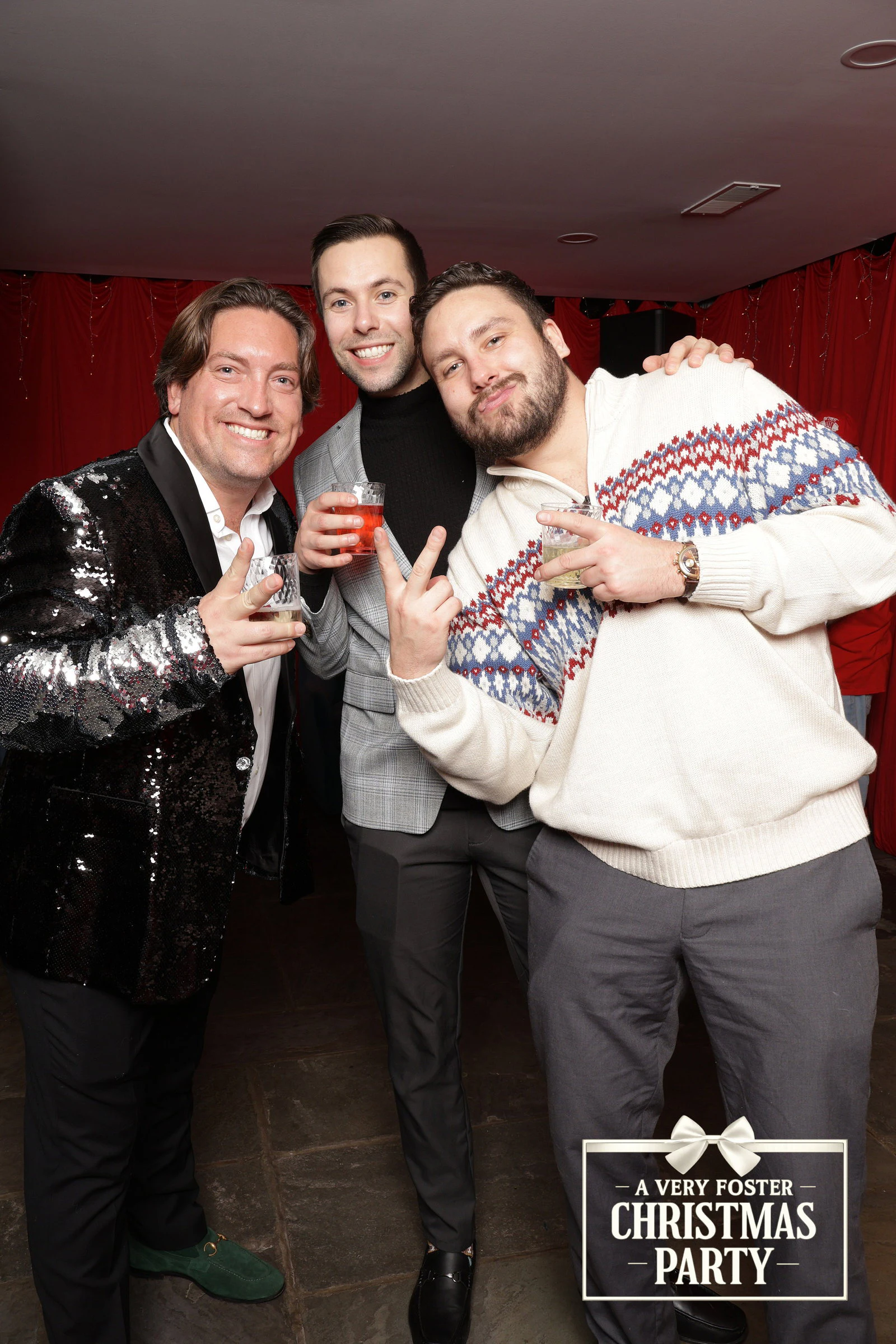 Three men, one in a black sequin jacket, one in a grey suit and one in a patterned cream sweater, raising their glasses in a lounge with red curtained walls at a private party