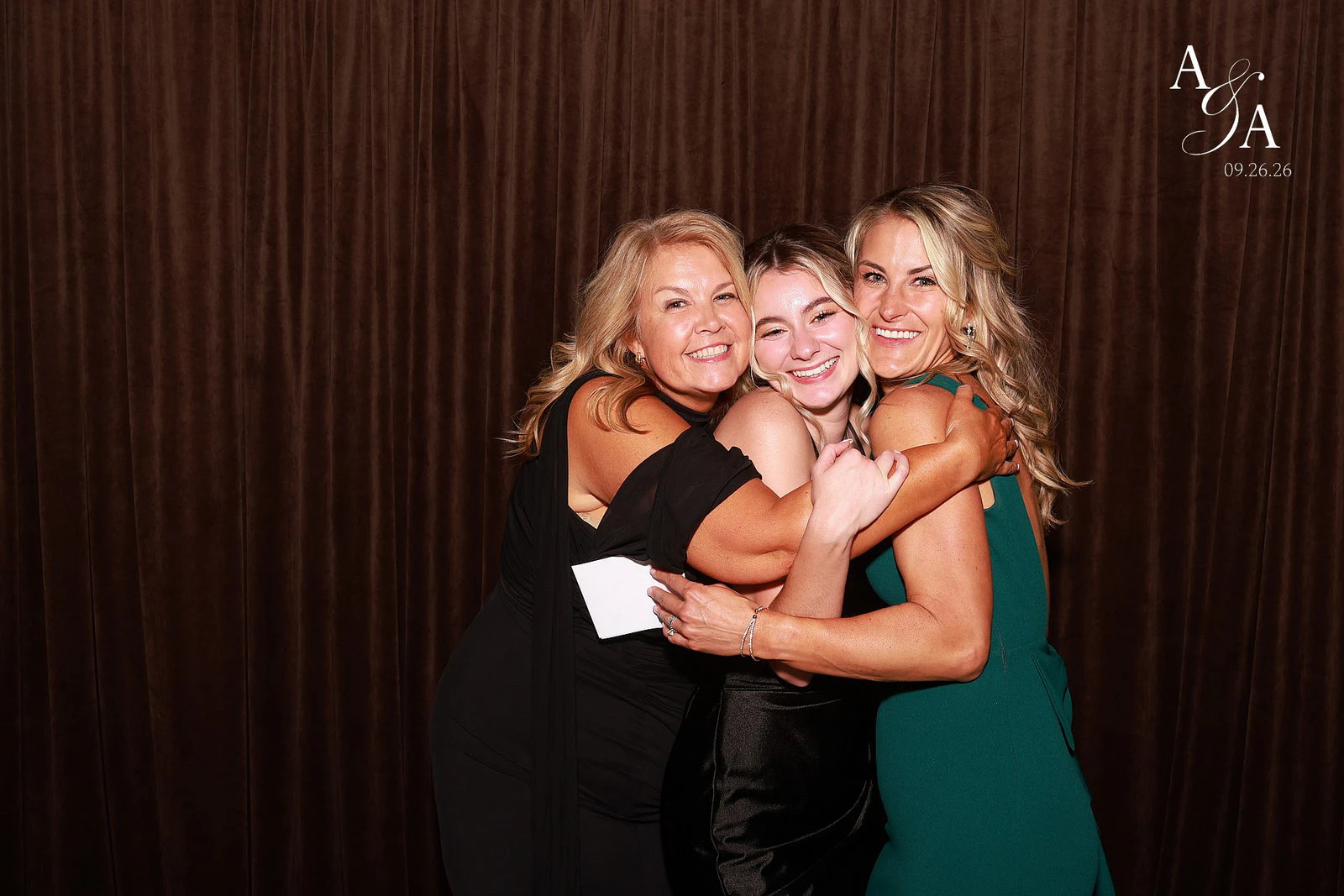 Three women in a tight hug, two in black and one in a green dress, all smiling at the camera at Alyssa + Alex