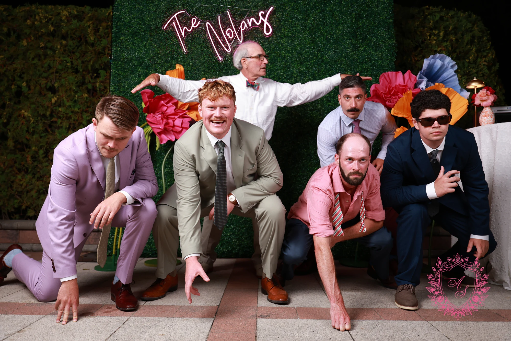 Six men in suits posing in front of the hedge wall, four crouched low and one behind with his arms out at Amanda + Stephen
