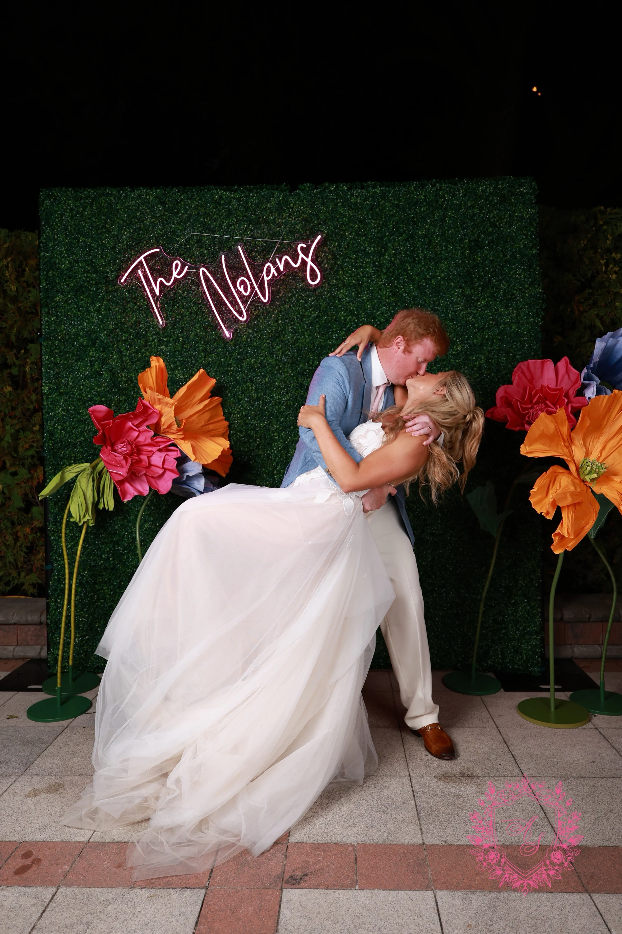 The man in the light-blue jacket dips the woman in the white gown for a kiss in front of the hedge wall at Amanda + Stephen