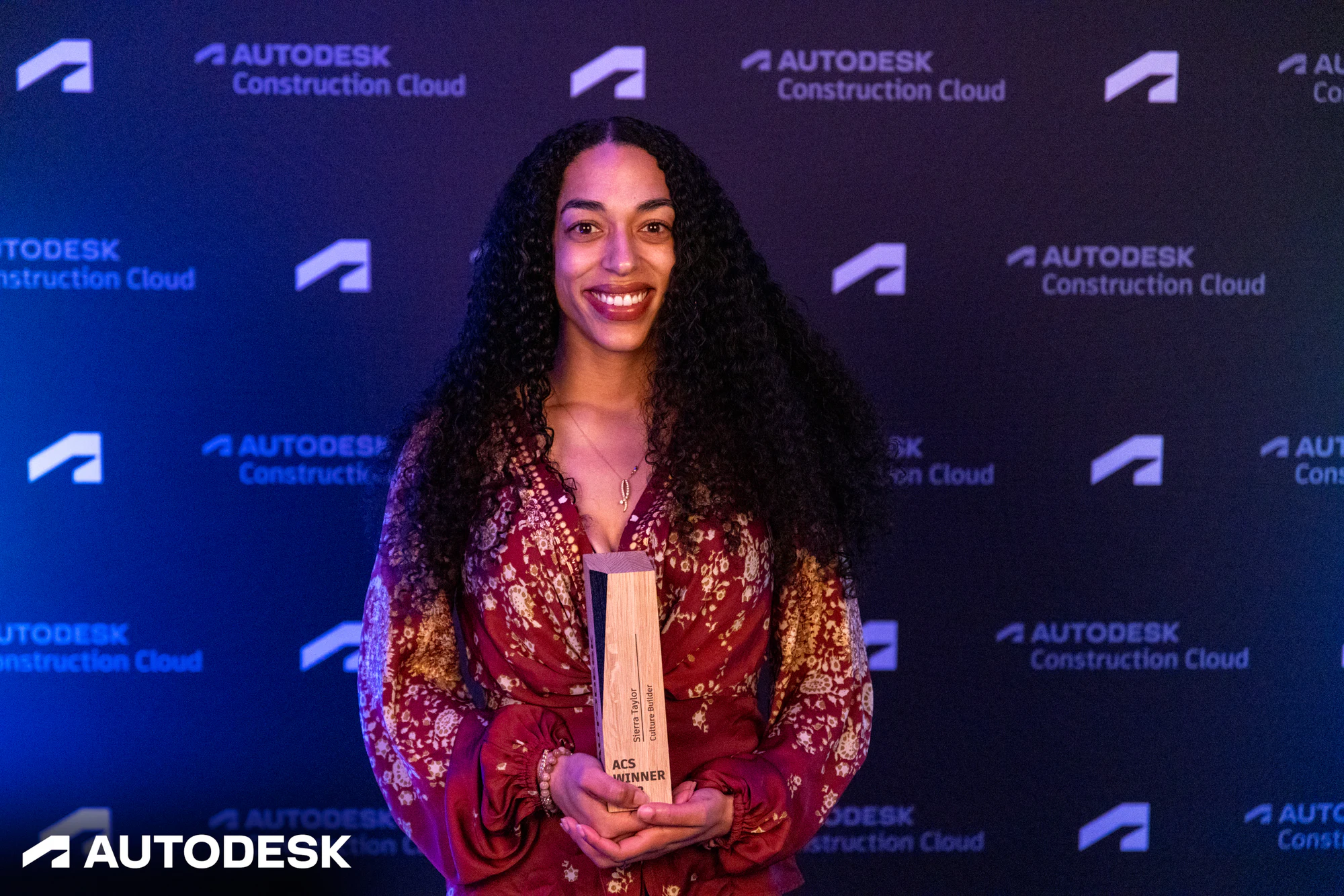 The winner smiling with her trophy at the backdrop at the Autodesk OTC Awards 2023