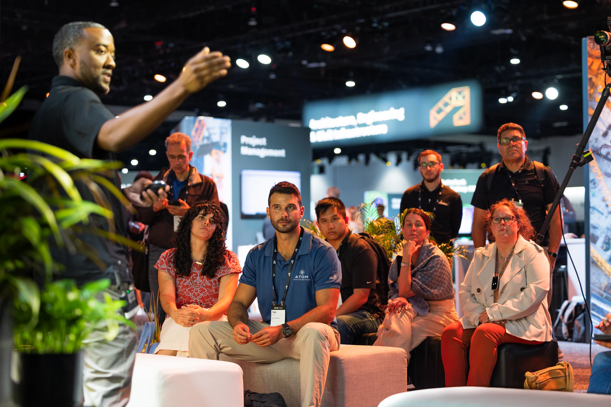 A speaker in a black polo talking with his hands to a seated audience in a lounge on the show floor at Autodesk University 2024
