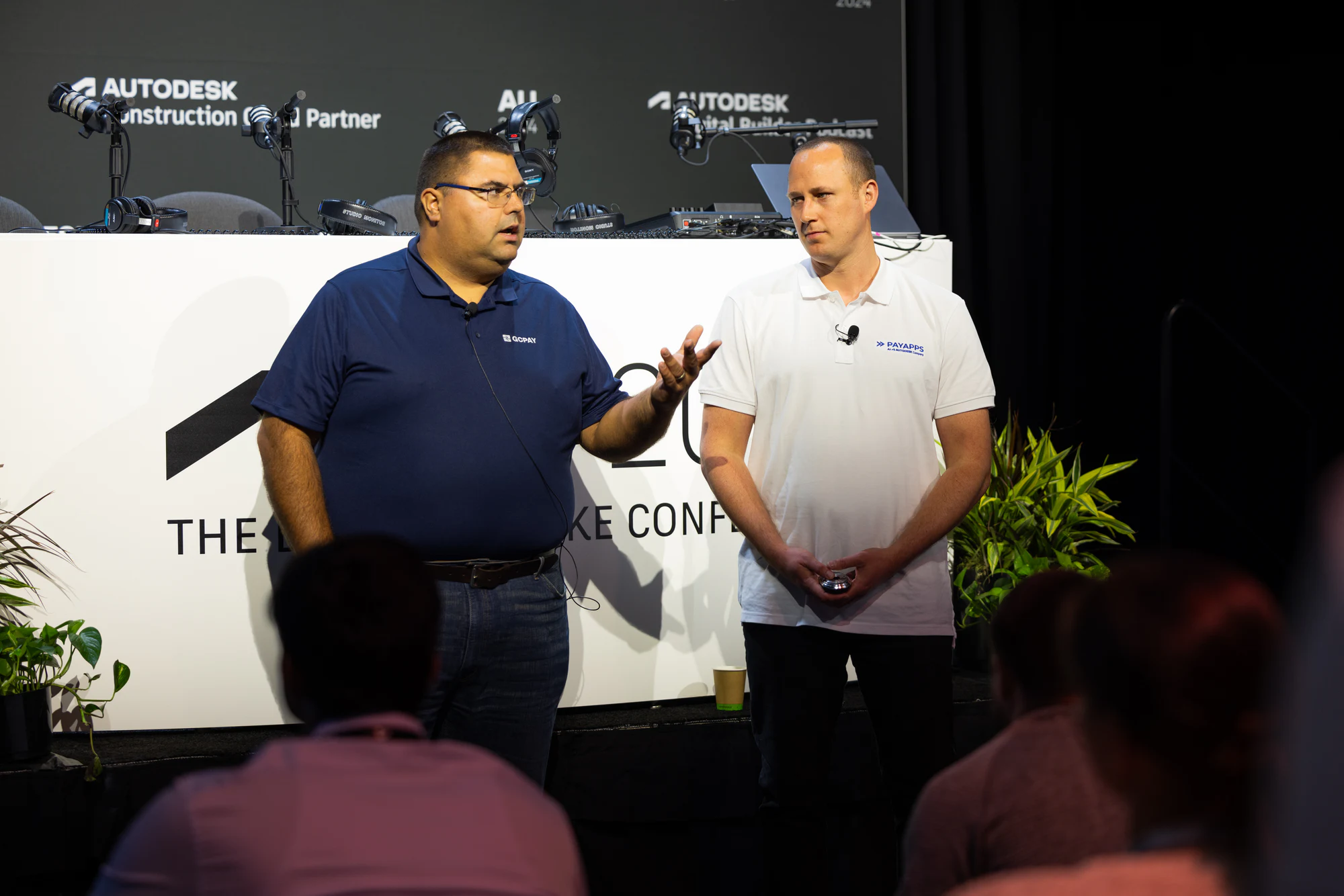 Two presenters in Autodesk Construction Cloud partner shirts on stage in front of the AU 2024 desk at Autodesk University 2024