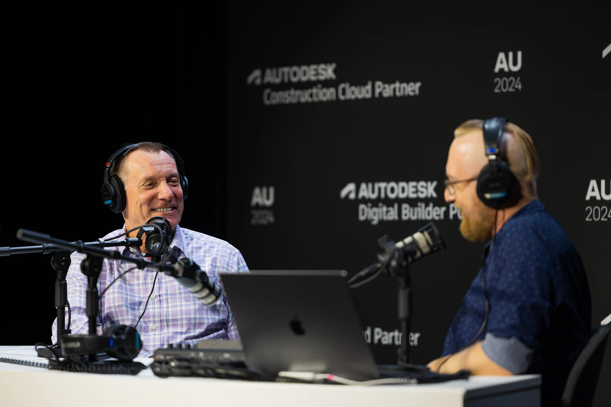 Two hosts in headphones laughing at the mics on the Digital Builder Podcast set at Autodesk University 2024