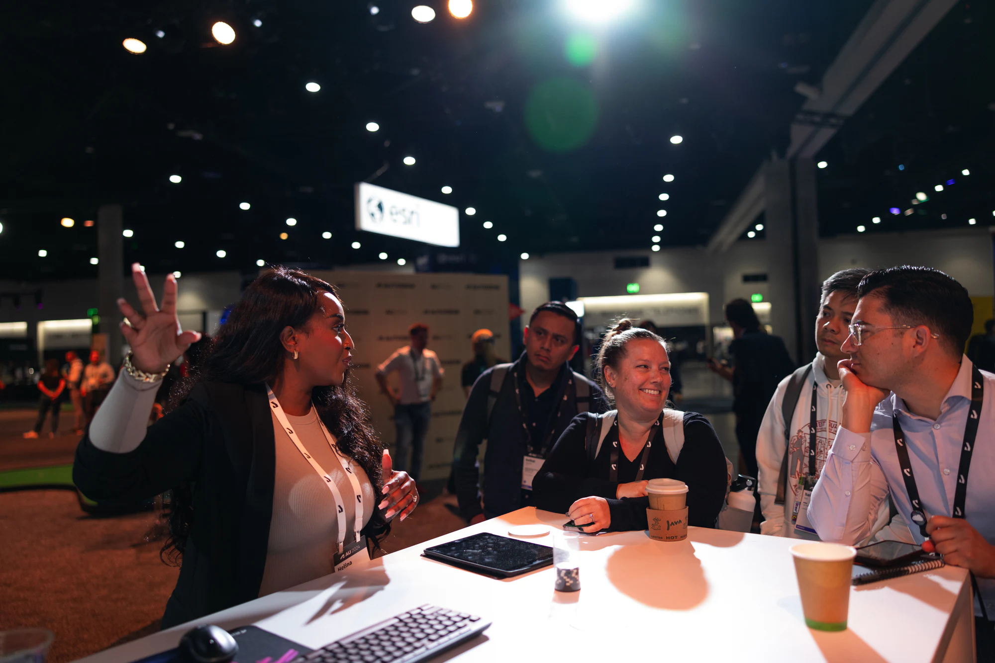 A product specialist talking with her hands at a demo table on the show floor, attendees listening over coffee at Autodesk University 2024