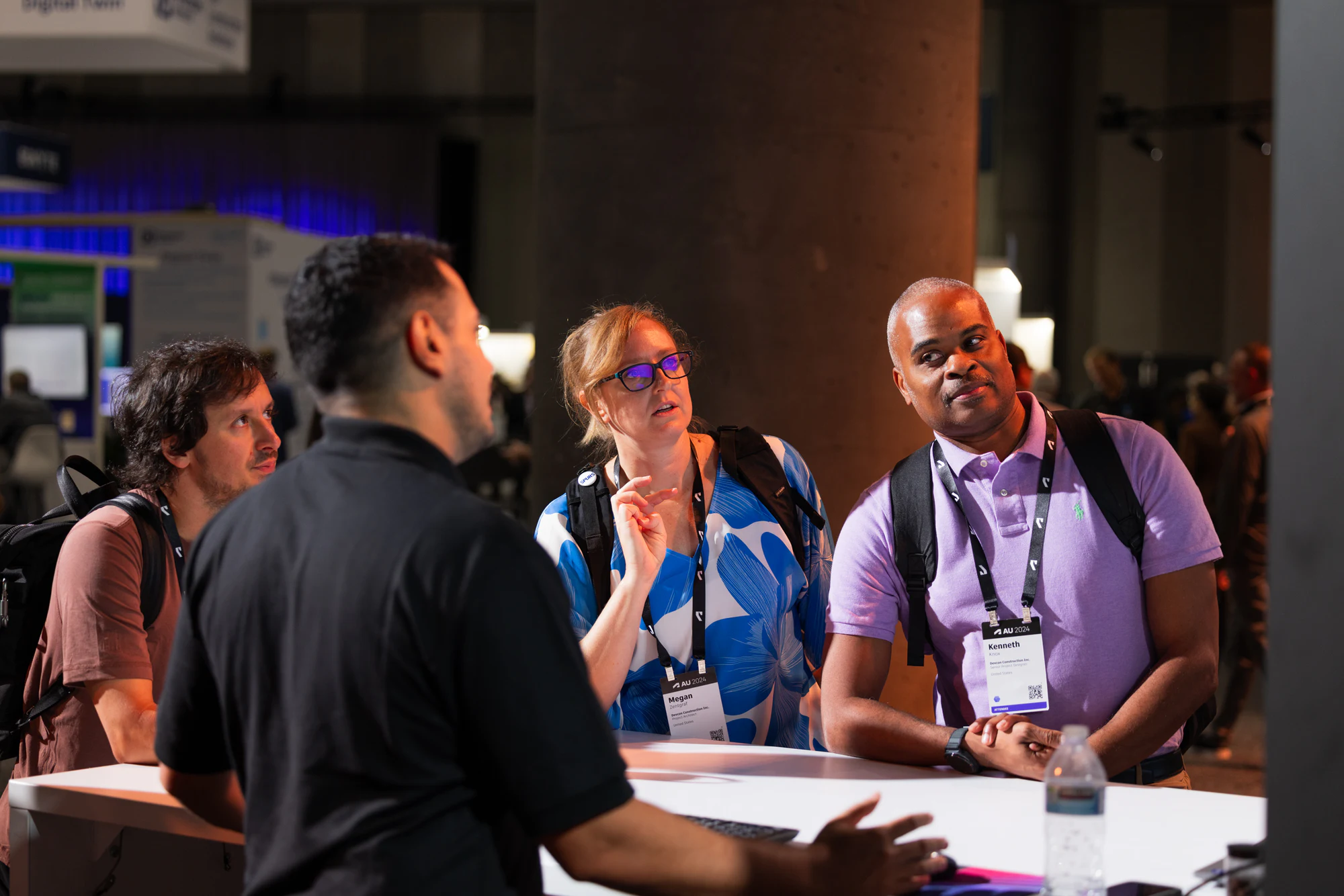 Three attendees in lanyards following a staffer's demo at a booth table at Autodesk University 2024