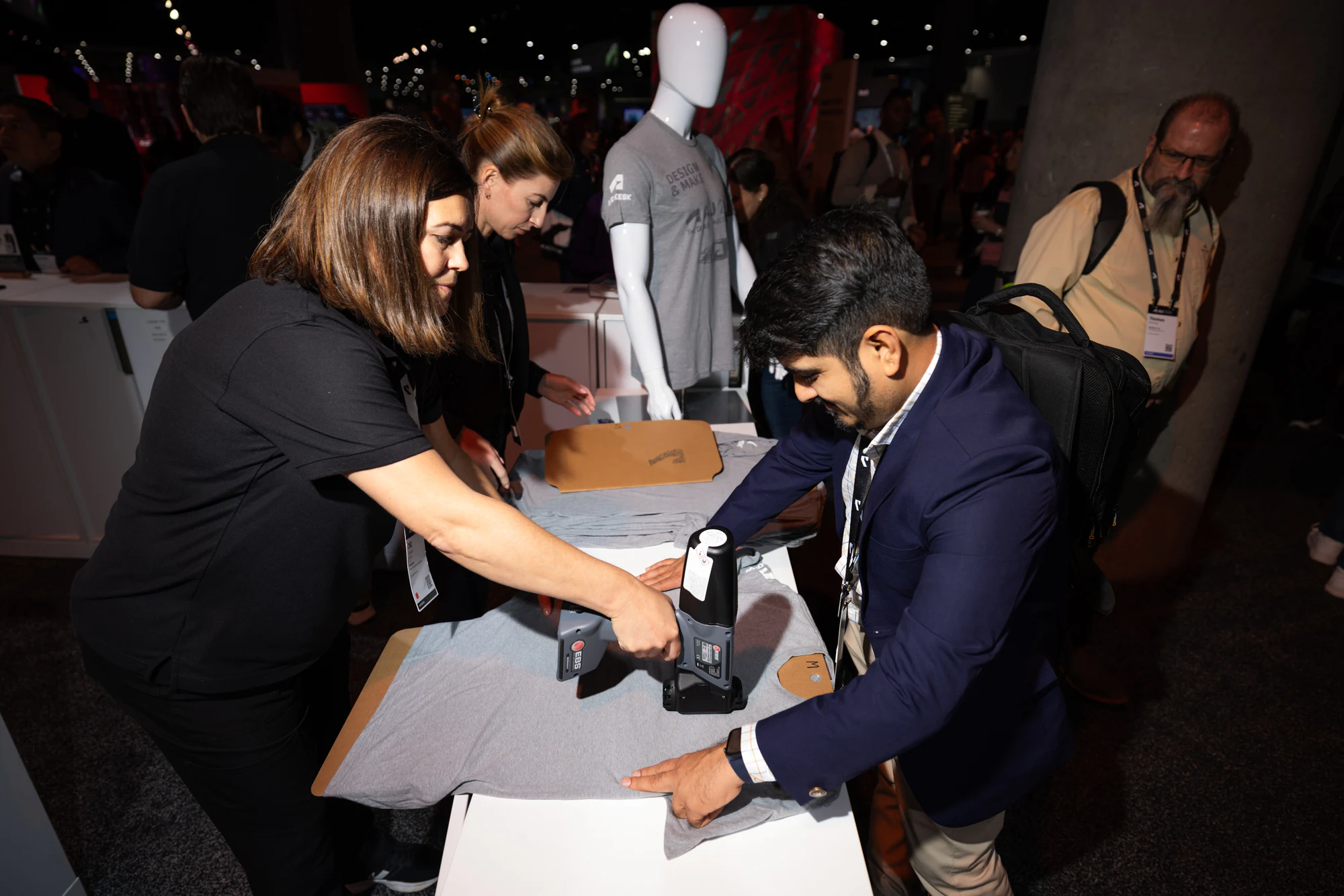 Attendees putting a design on a T-shirt at a merch station on the show floor, a mannequin in a finished shirt behind them at Autodesk University 2024