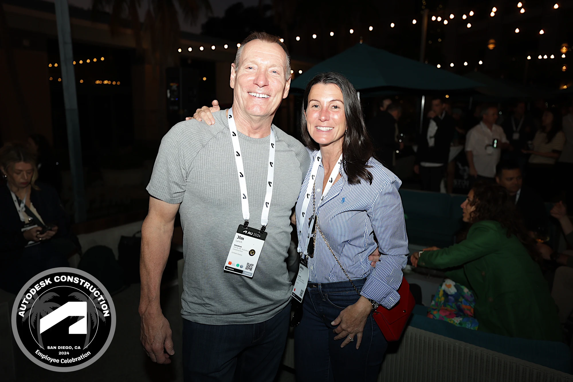 Two coworkers in lanyards arm in arm under the string lights at Autodesk University 2024