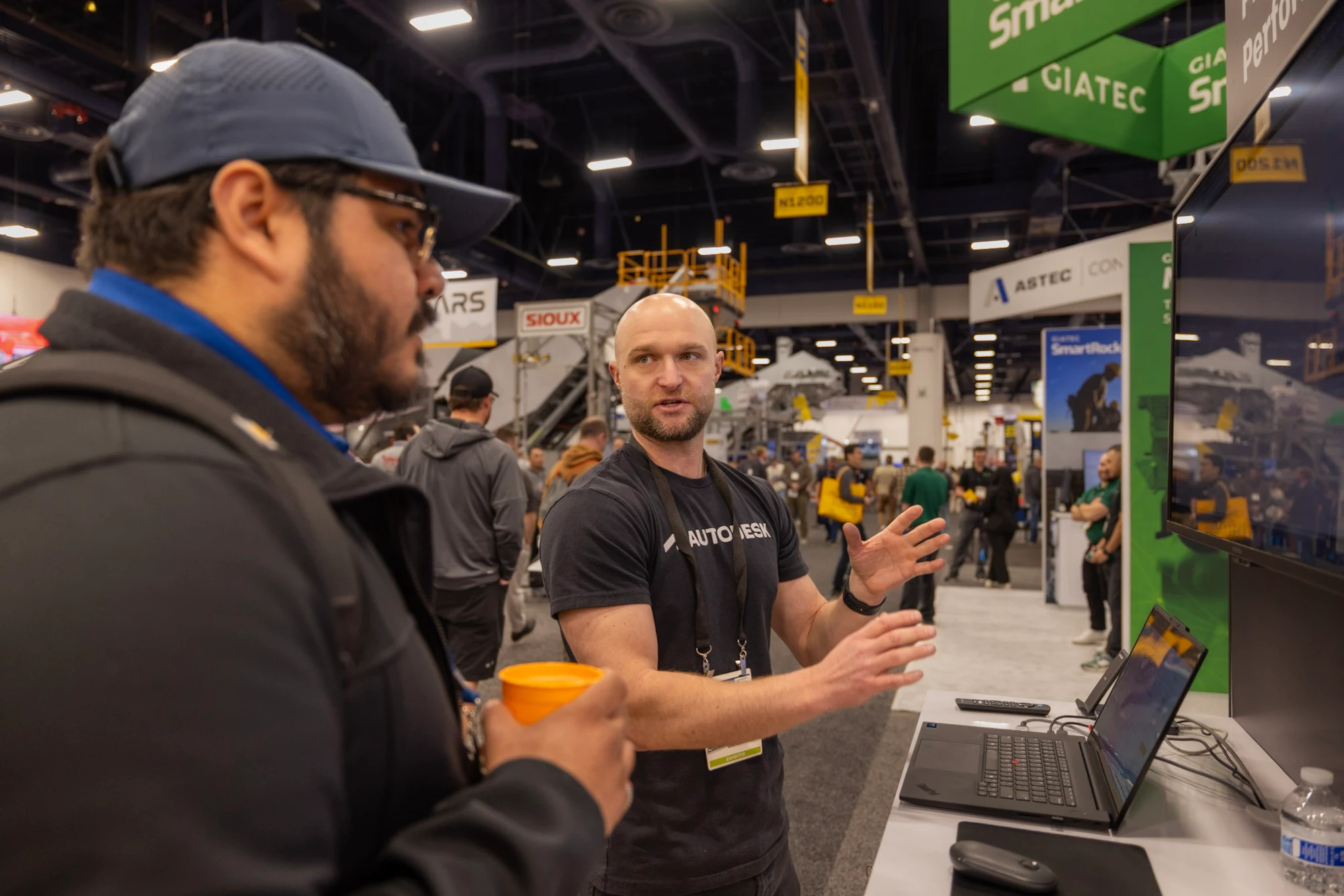 The team member explaining the demo while the visitor holds his drink at Autodesk at World of Concrete 2026