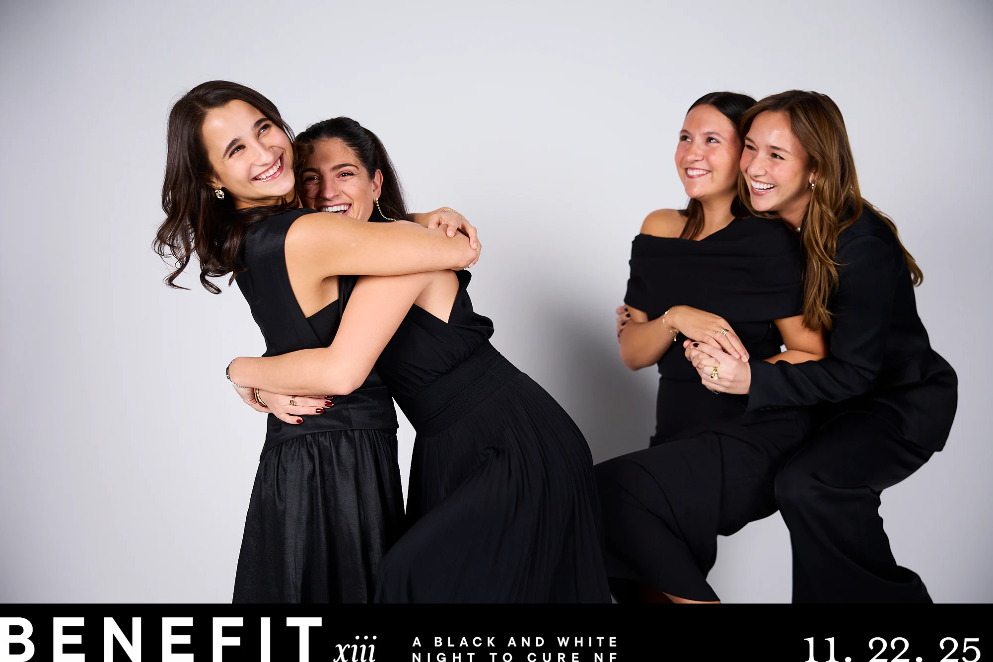 Four friends in black dresses hugging and laughing against a white backdrop at beNeFit XIII