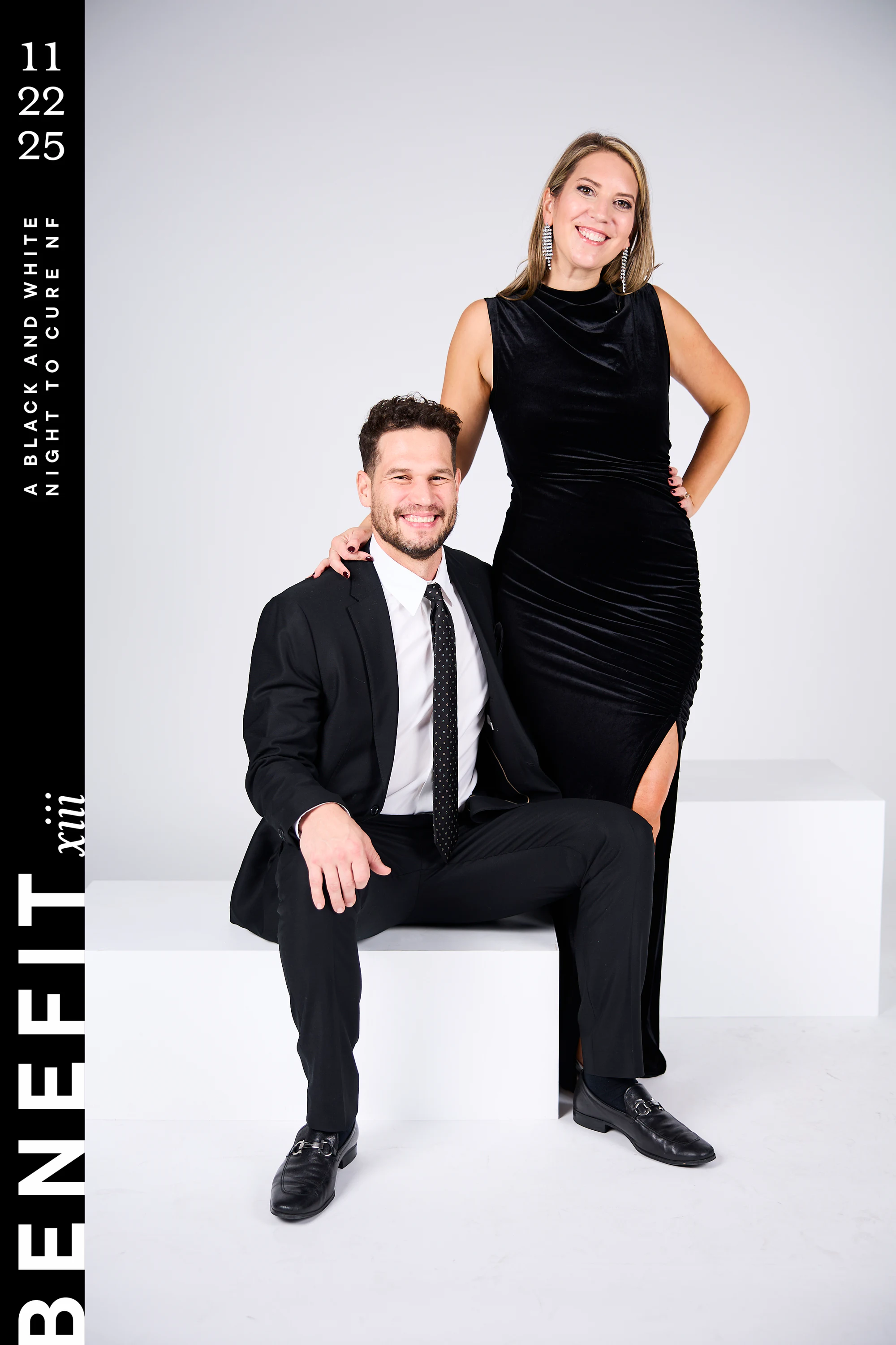 A couple in black, him seated on a white block, against the white backdrop at beNeFit XIII
