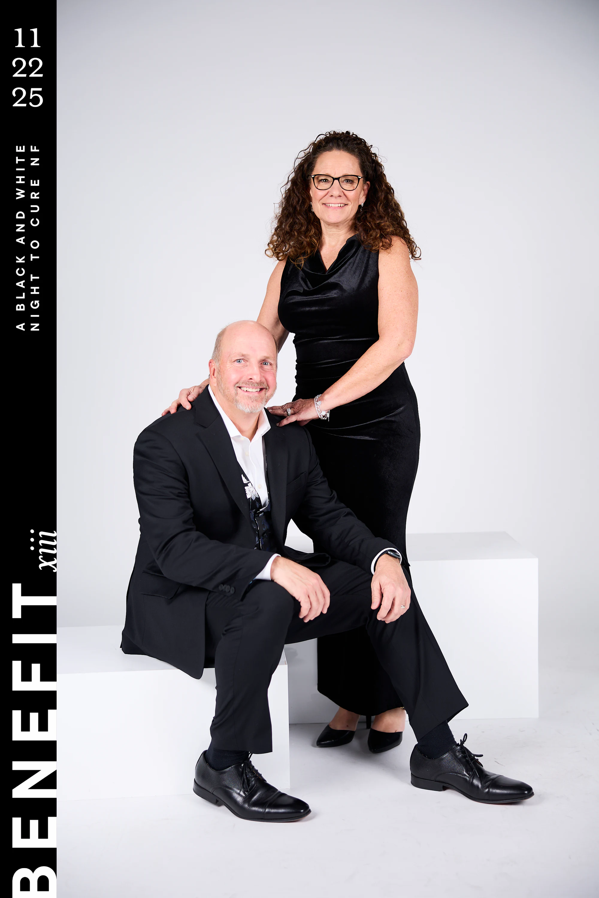 A couple in black, him seated on a white block, her standing behind him in a velvet dress and glasses at beNeFit XIII
