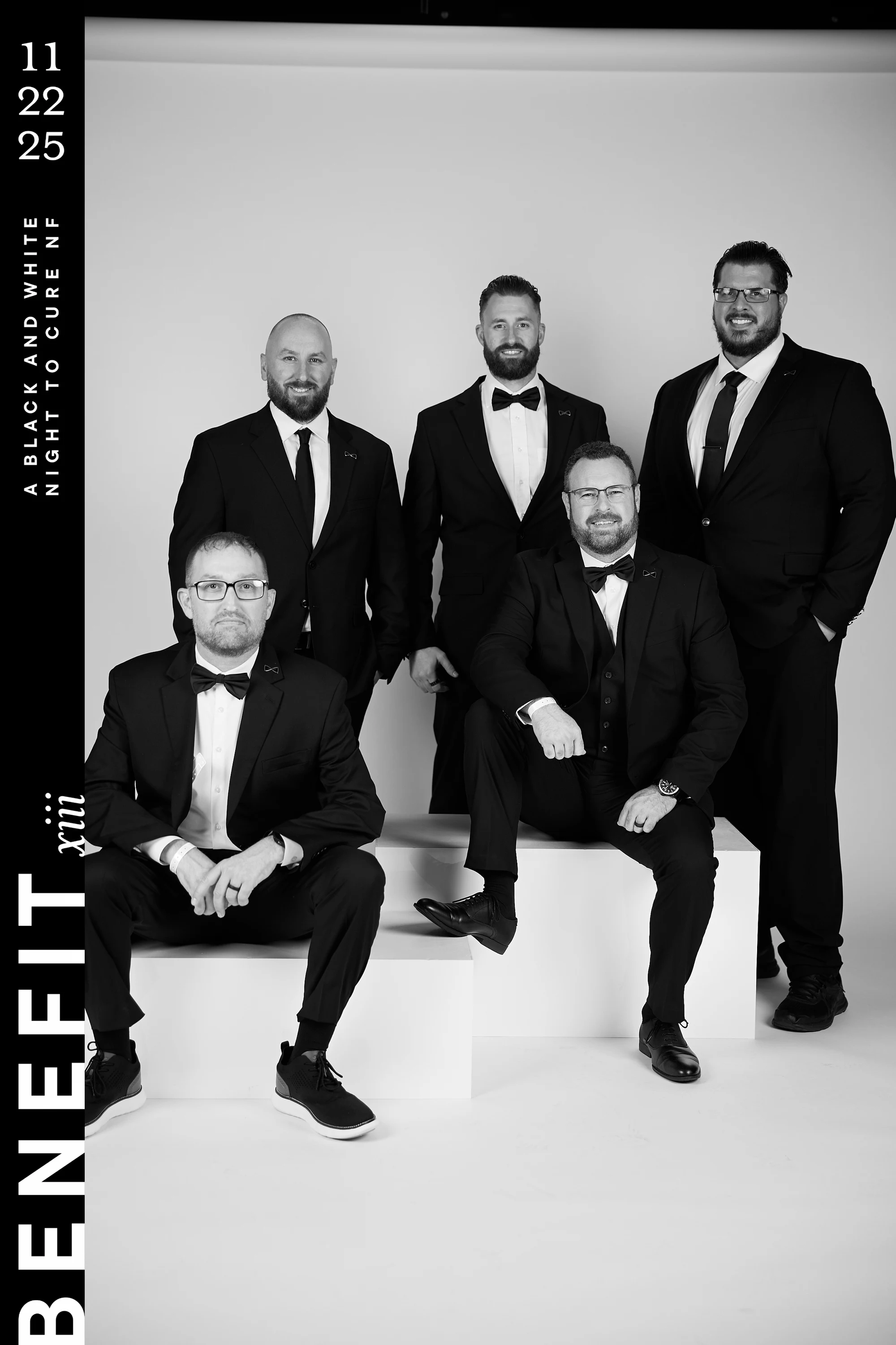 Black and white portrait of five men in black suits and bow ties posed on white blocks at beNeFit XIII