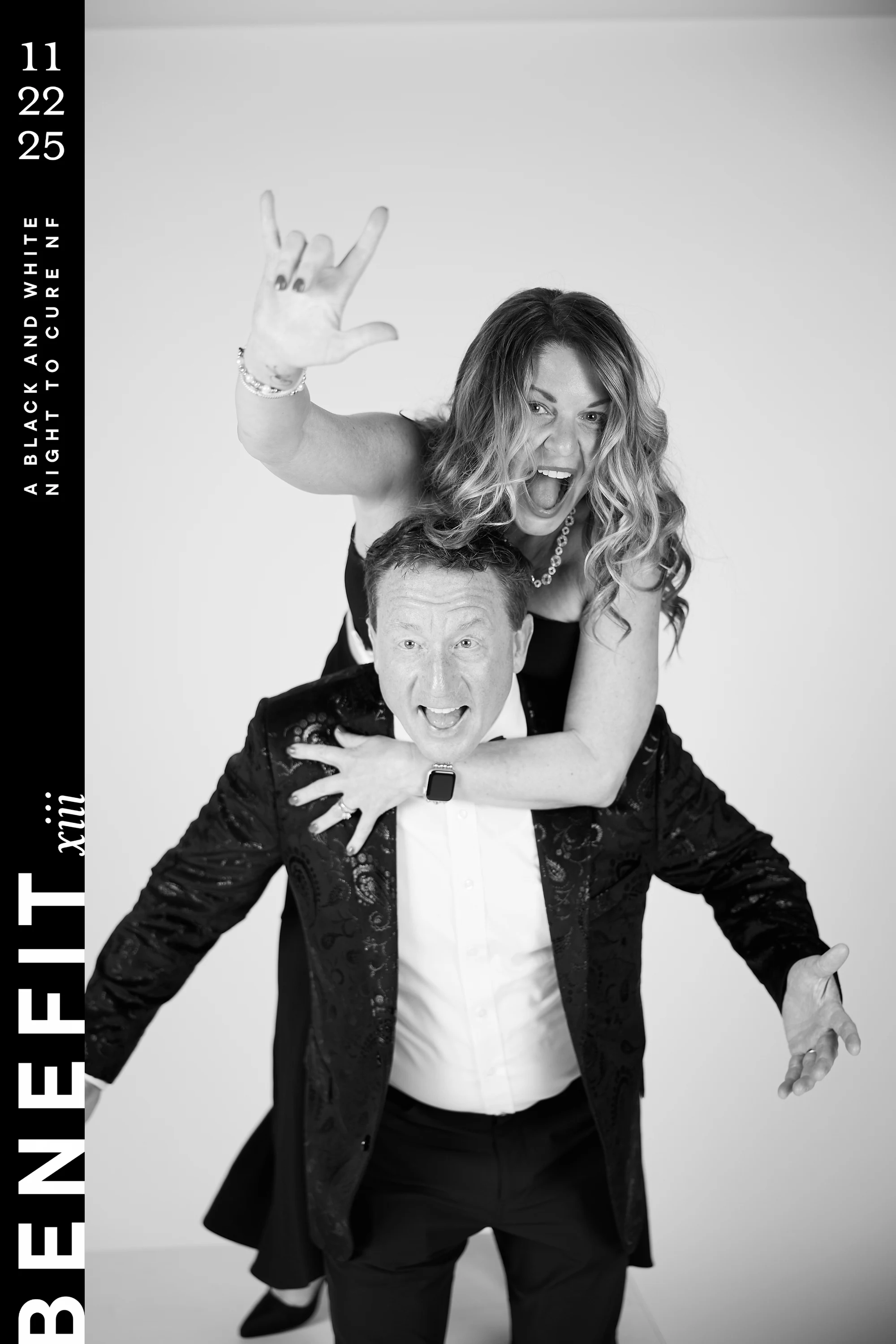 Black and white portrait of a woman riding piggyback on a man, throwing a rock-on sign as they both shout at beNeFit XIII