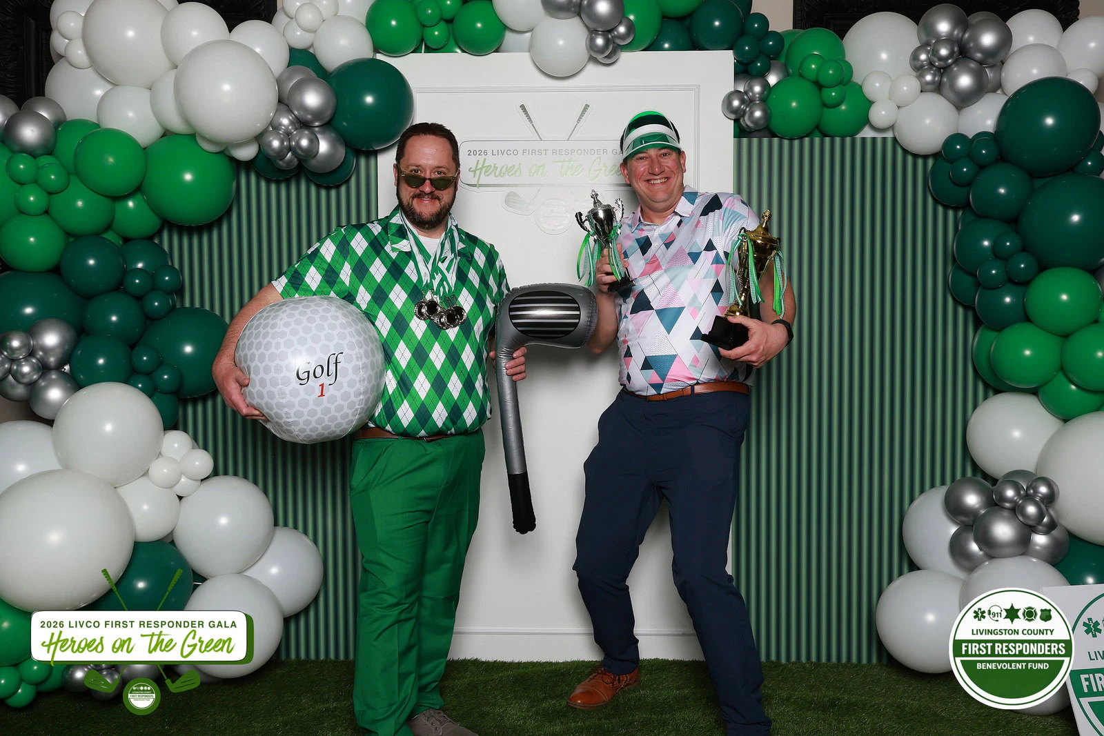 Two men in loud golf shirts, one hugging a giant golf ball and an inflatable club, the other holding two trophies at Benevolent Fund Gala