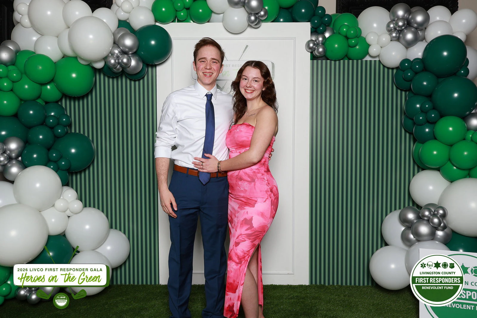 A man in a white shirt and blue tie and a woman in a pink patterned dress standing close in front of a green and white balloon arch at Benevolent Fund Gala
