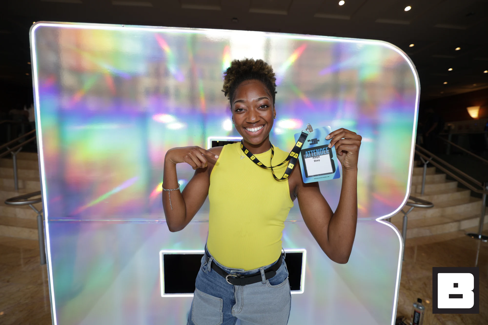 A guest in a yellow top holding up her badge in front of the iridescent B at Black Tech Week 2025