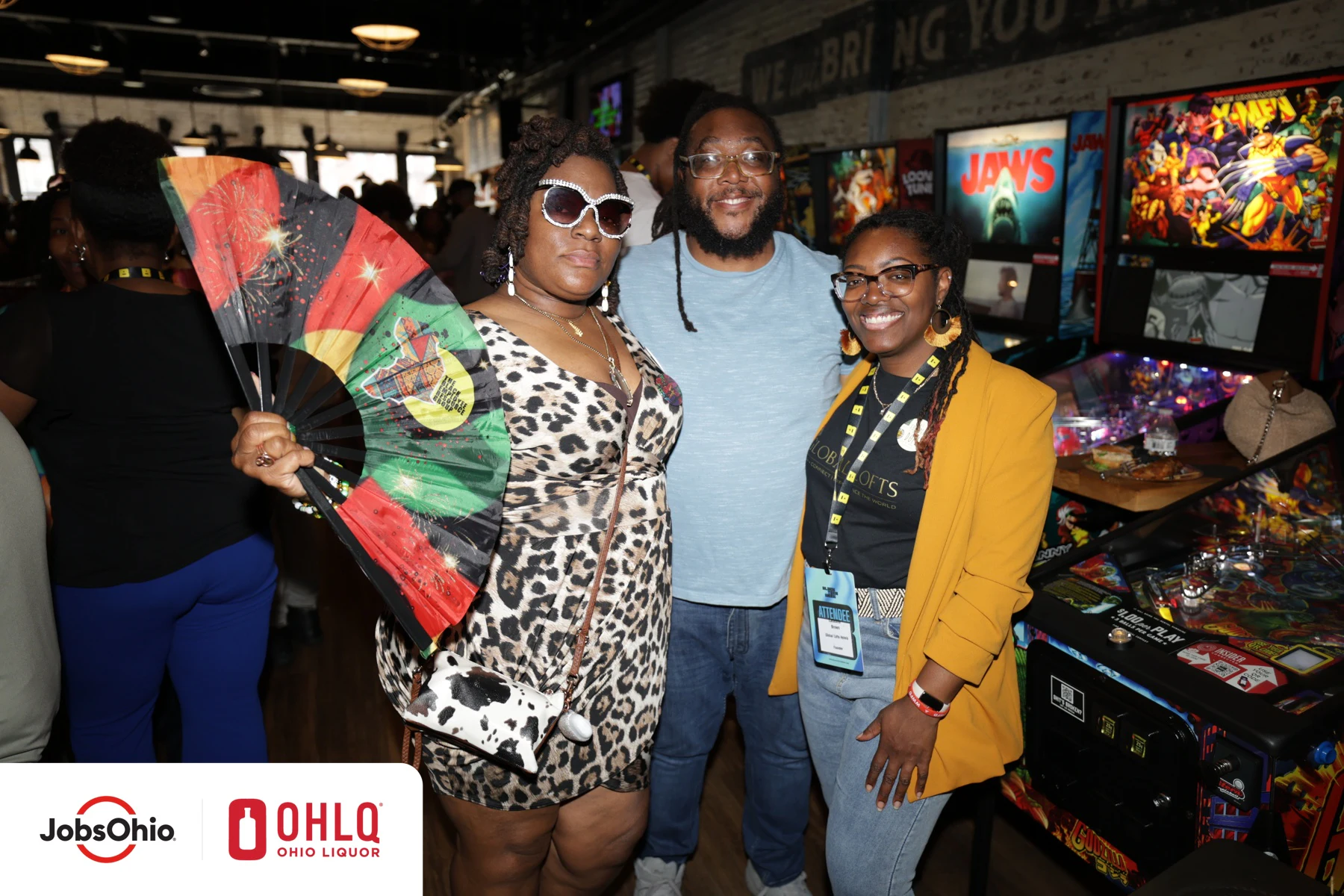 Three guests by the pinball machines, one holding a large fan, at the closing reception at Black Tech Week 2025
