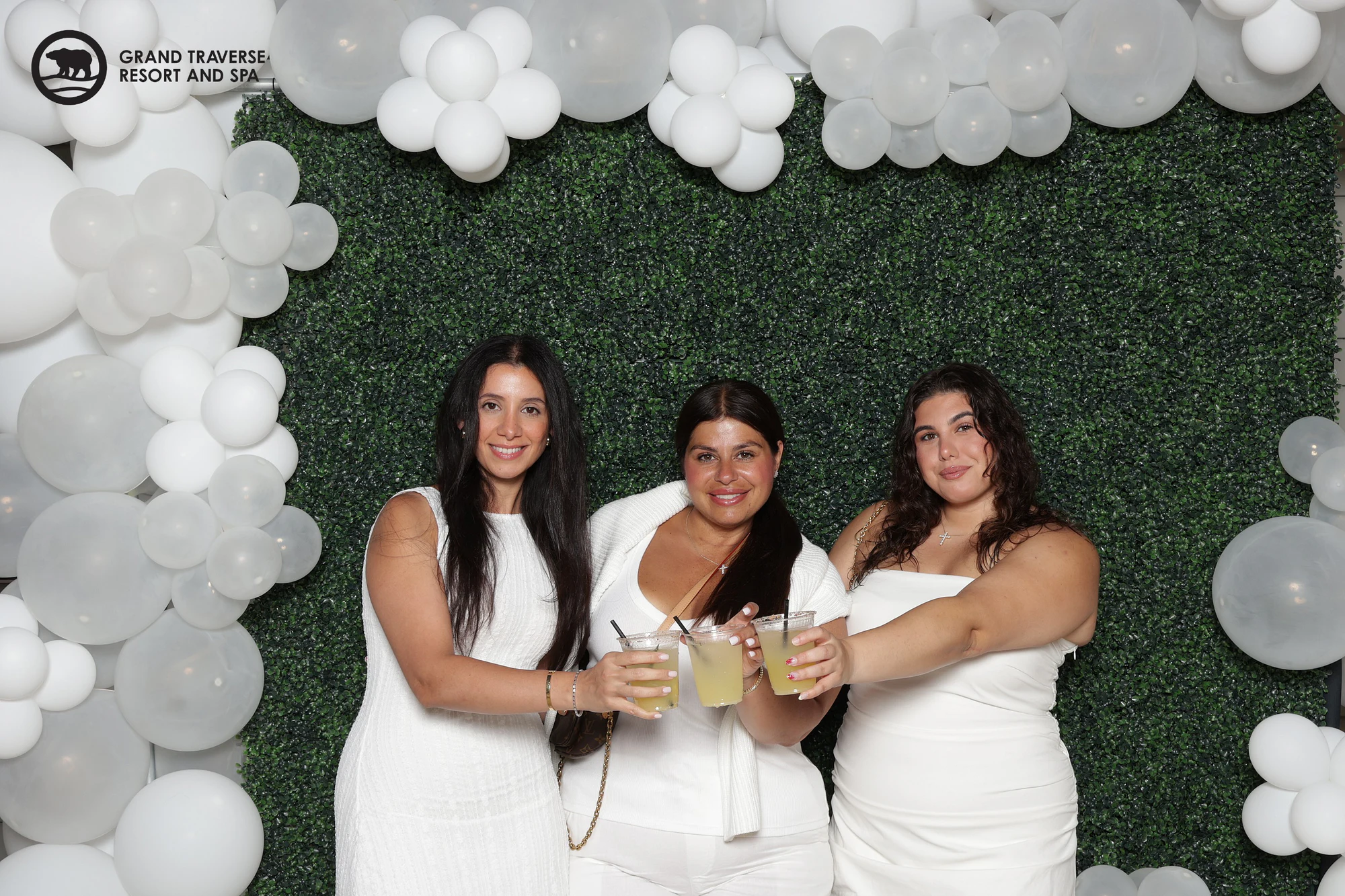 Three women in white dresses clinking their drinks together in front of the hedge wall at Blanco Night at Grand Traverse Resort