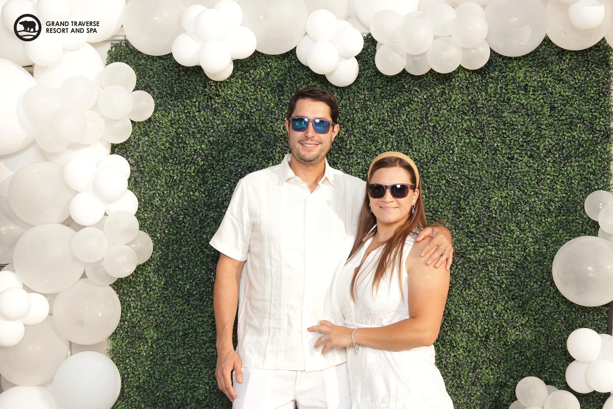 A man in a white linen shirt and sunglasses with his arm around a woman in a white dress, white balloons around the hedge wall behind them at Blanco Night at Grand Traverse Resort