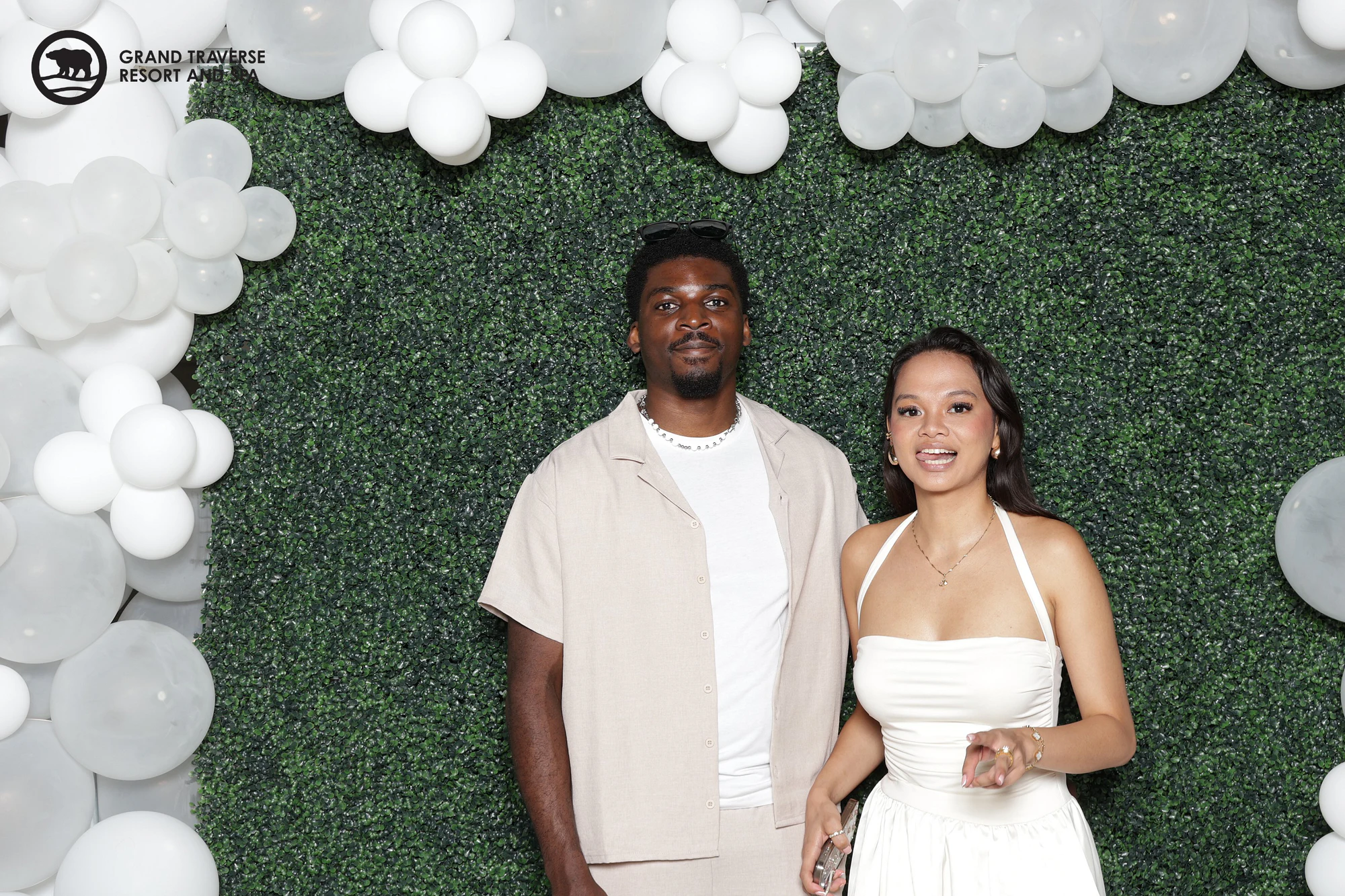 A man in a tan overshirt and a woman in a white halter dress standing side by side in front of the hedge wall at Blanco Night at Grand Traverse Resort