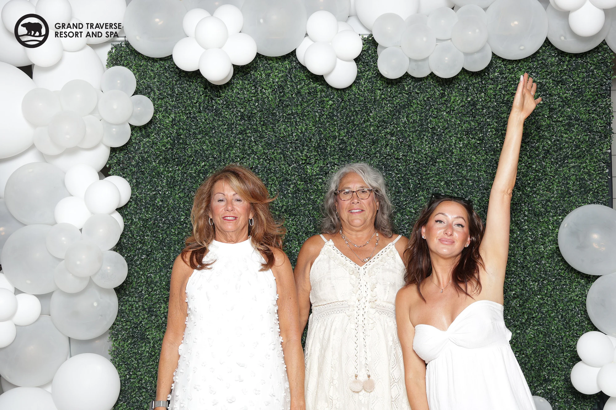 Three women in white dresses, one with her arm stretched straight up, in front of the hedge wall at Blanco Night at Grand Traverse Resort