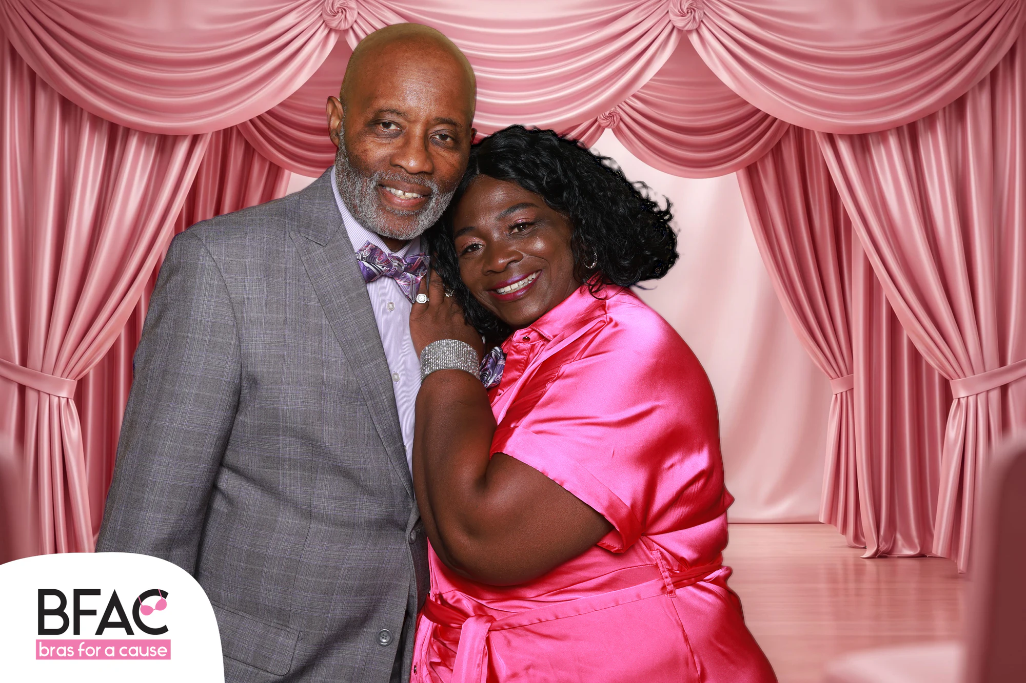 A man in a grey suit and a woman in pink satin hugging in front of pink curtains at Bras for a Cause 2026
