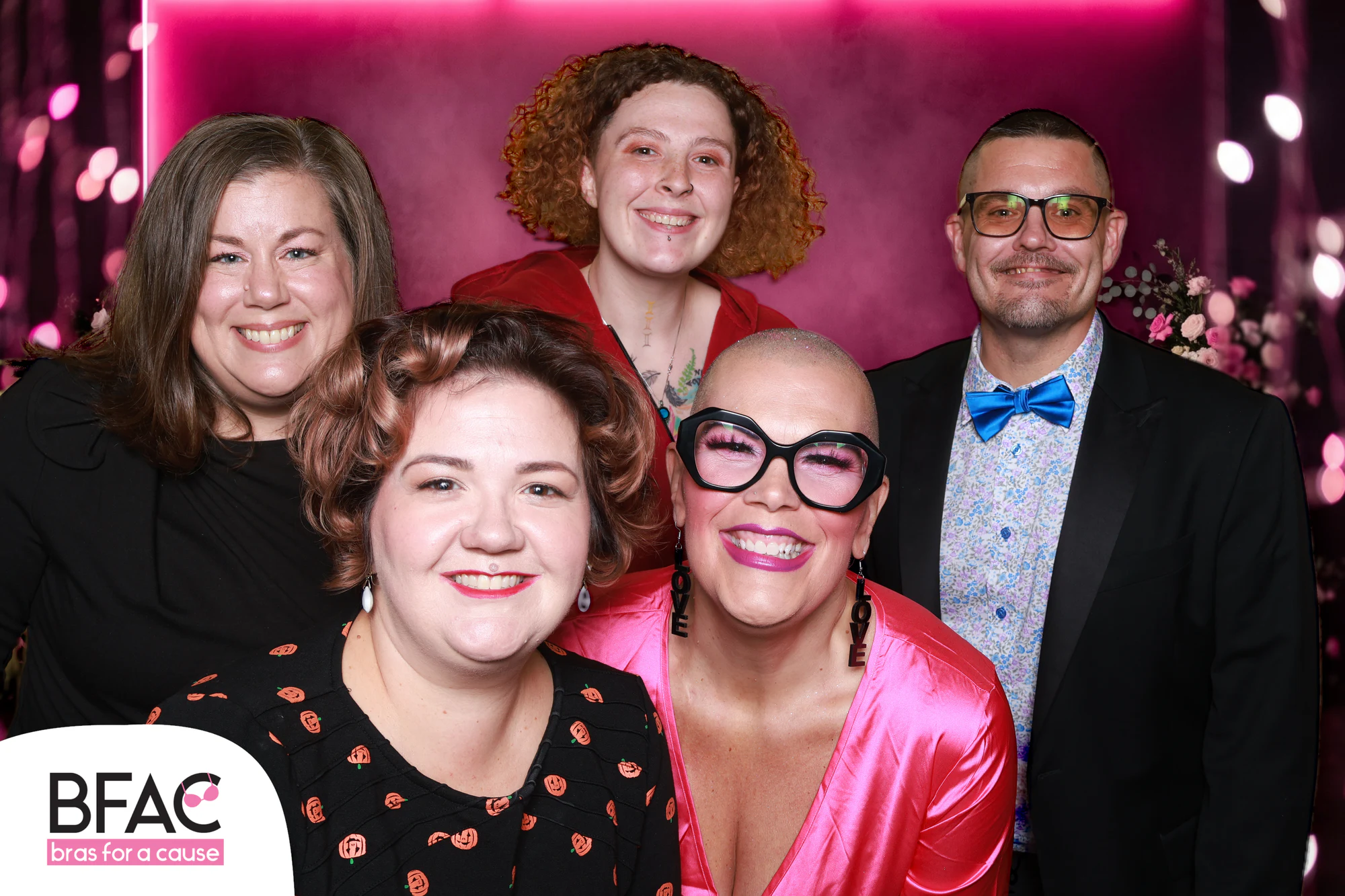Six friends crowded together on the pink stage, one woman in pink satin and big glasses at Bras for a Cause 2026