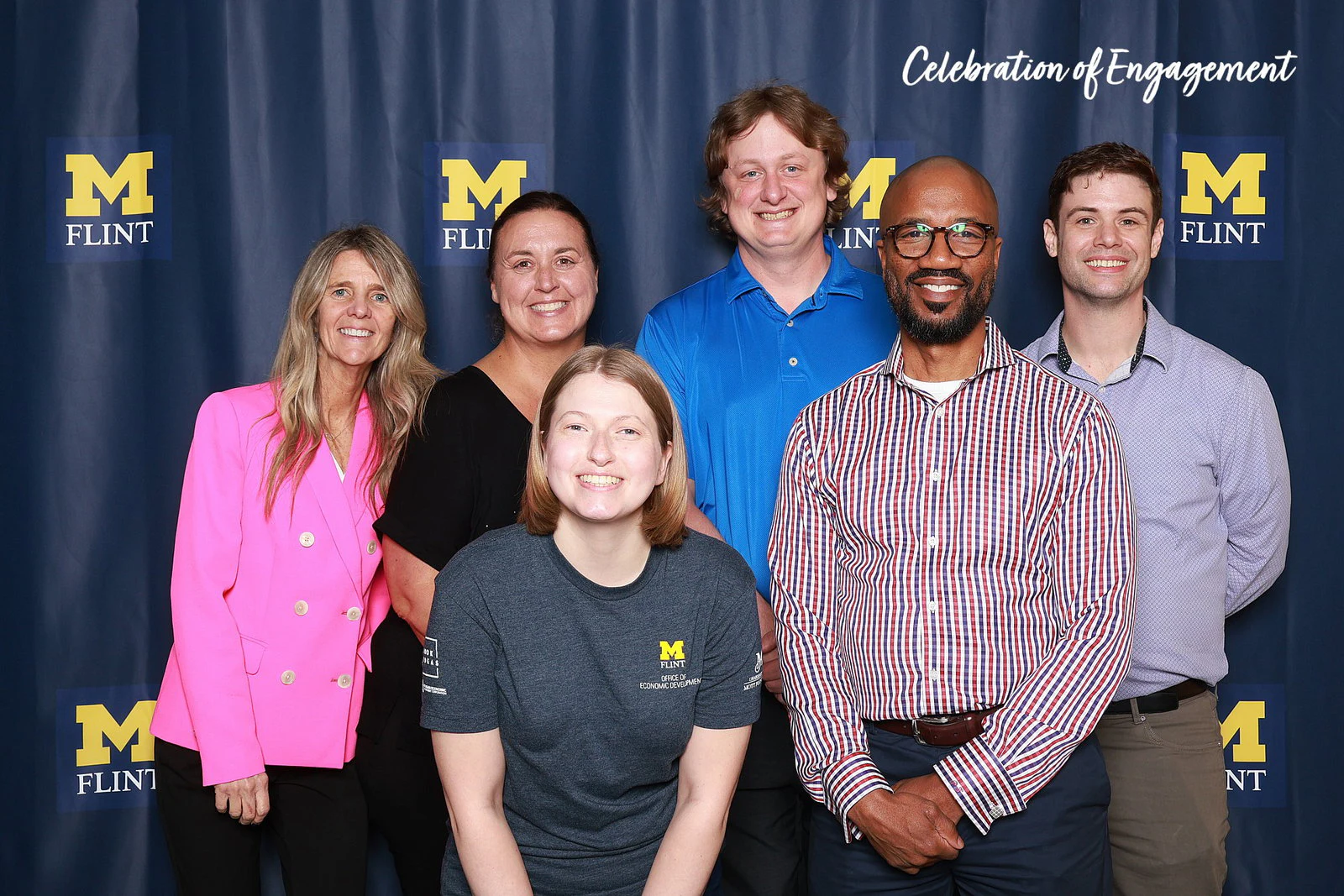 Six colleagues, one in a pink blazer, standing in two rows in front of a navy drape printed with a repeating logo at Celebration of Engagement