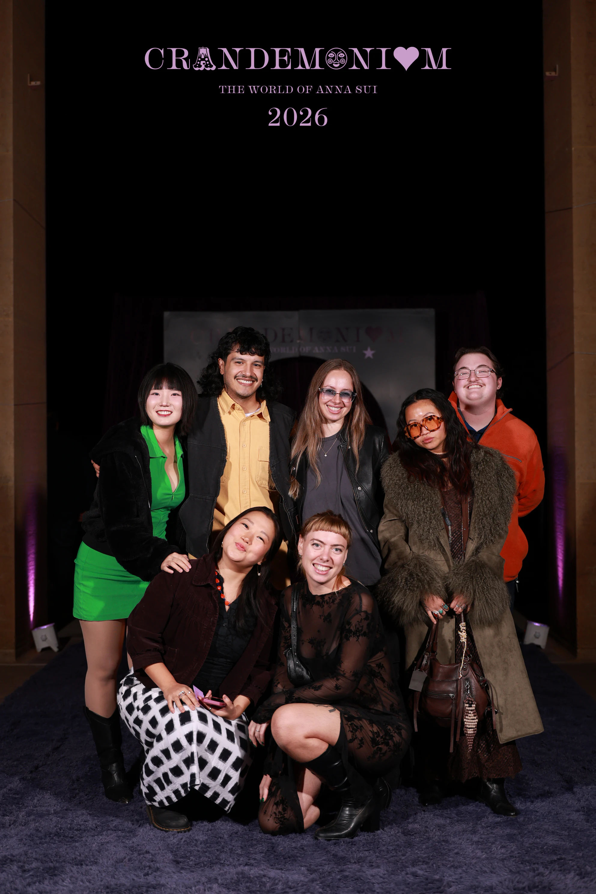Nine guests on the purple carpet in front of the arch, some kneeling in front at Crandemonium 2026