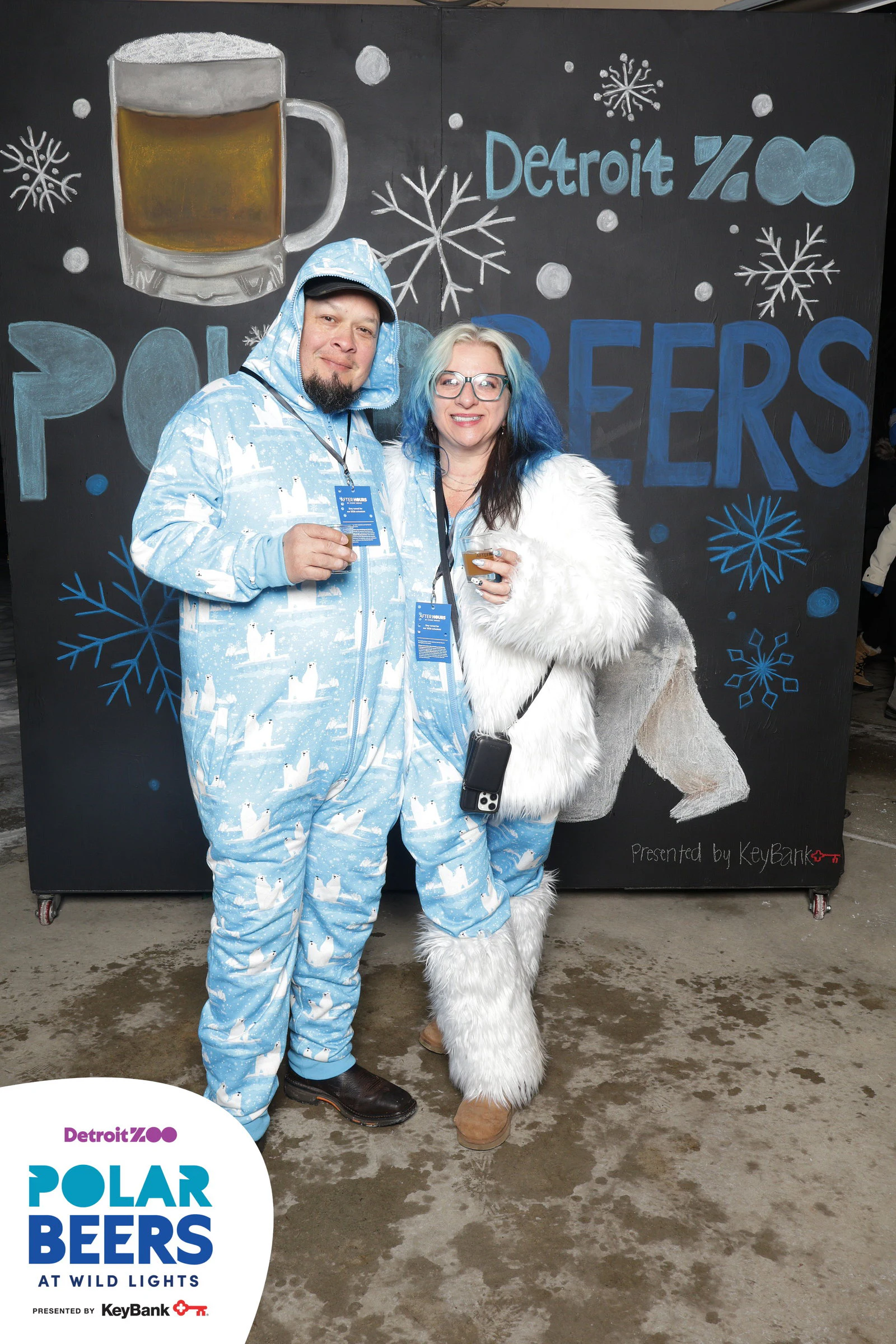 A man in a pale blue polar bear onesie and a woman in a white fur coat and fur boots in front of a hand-painted Polar Beers wall at the Detroit Zoo