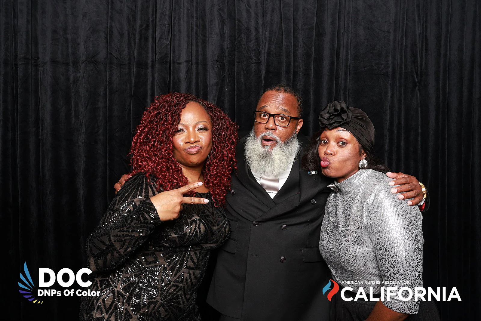 A man in a black suit between two women in black and silver, peace signs up at DNPs of Color