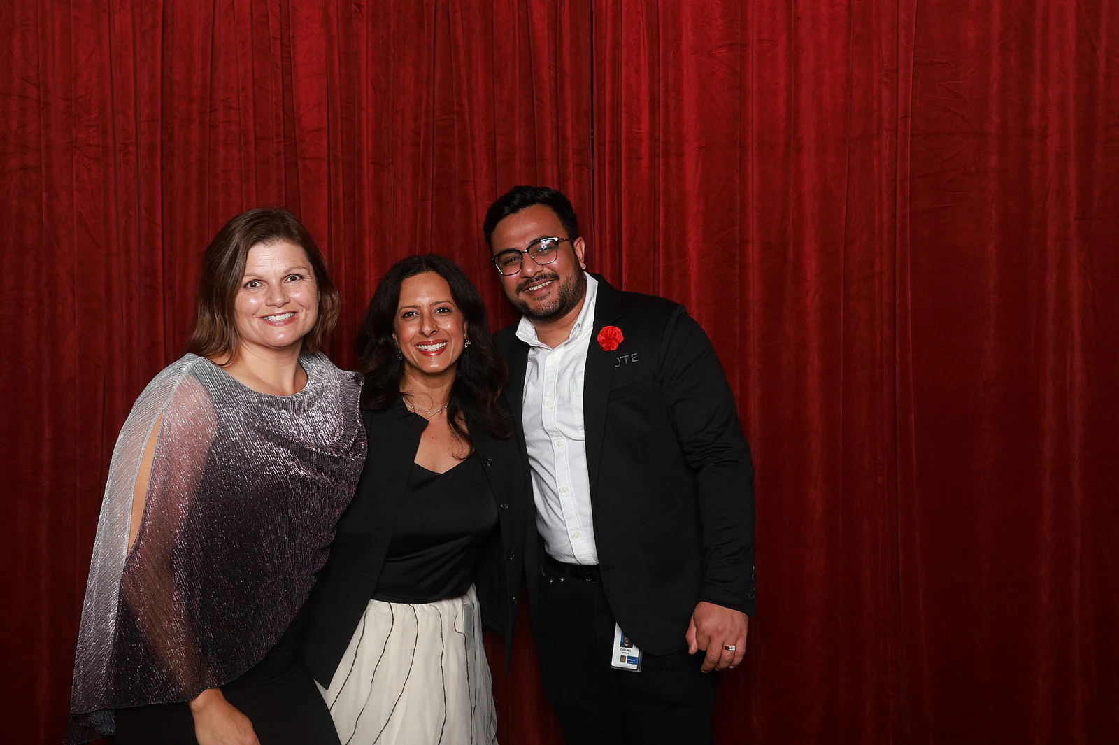 A woman in a silver shimmer wrap, a woman in black and a man in a black jacket with a red flower on the lapel at DTE Gala
