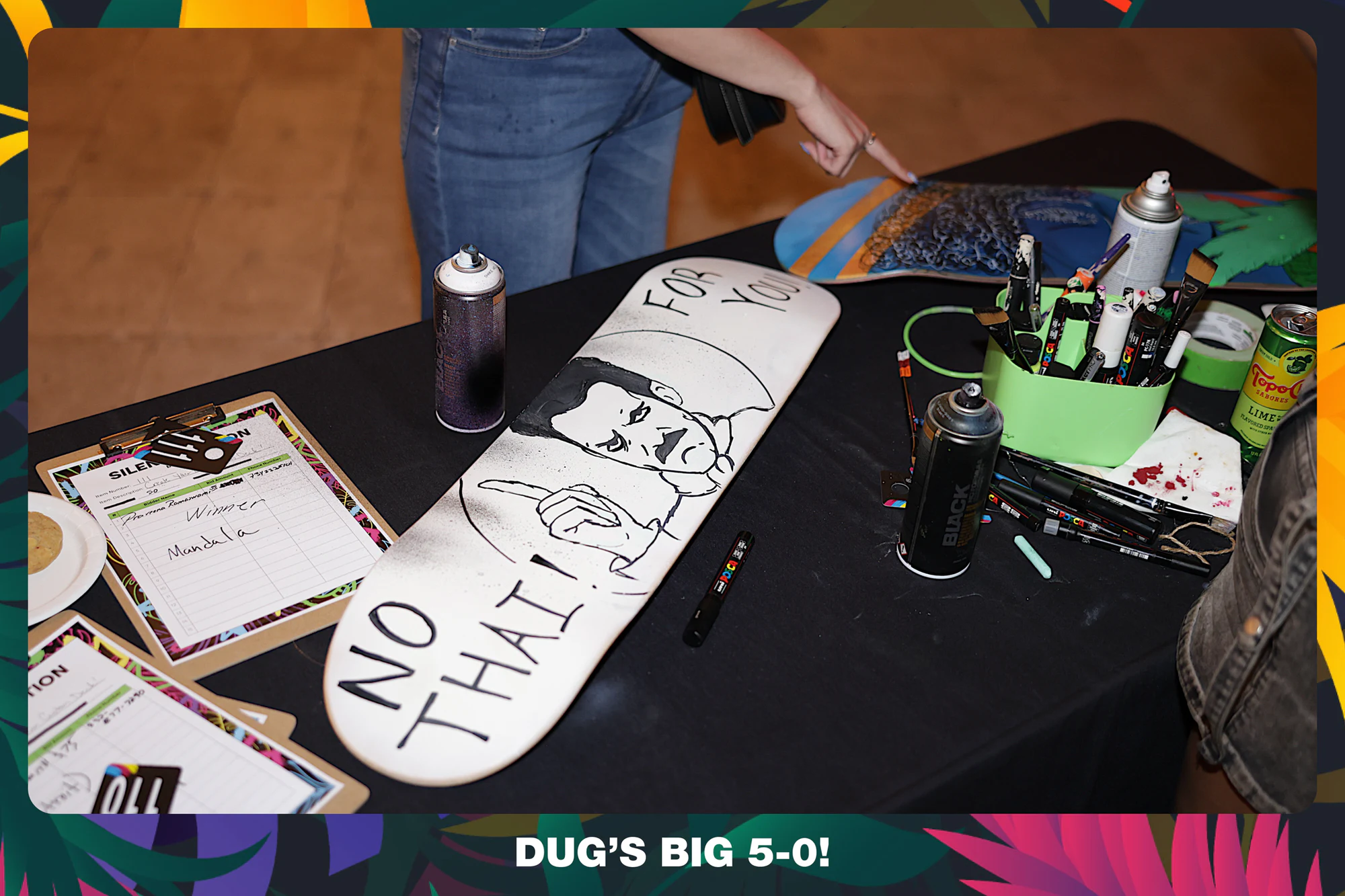 A hand-drawn skate deck on the art table with spray cans and markers at Dug's Big 5-0