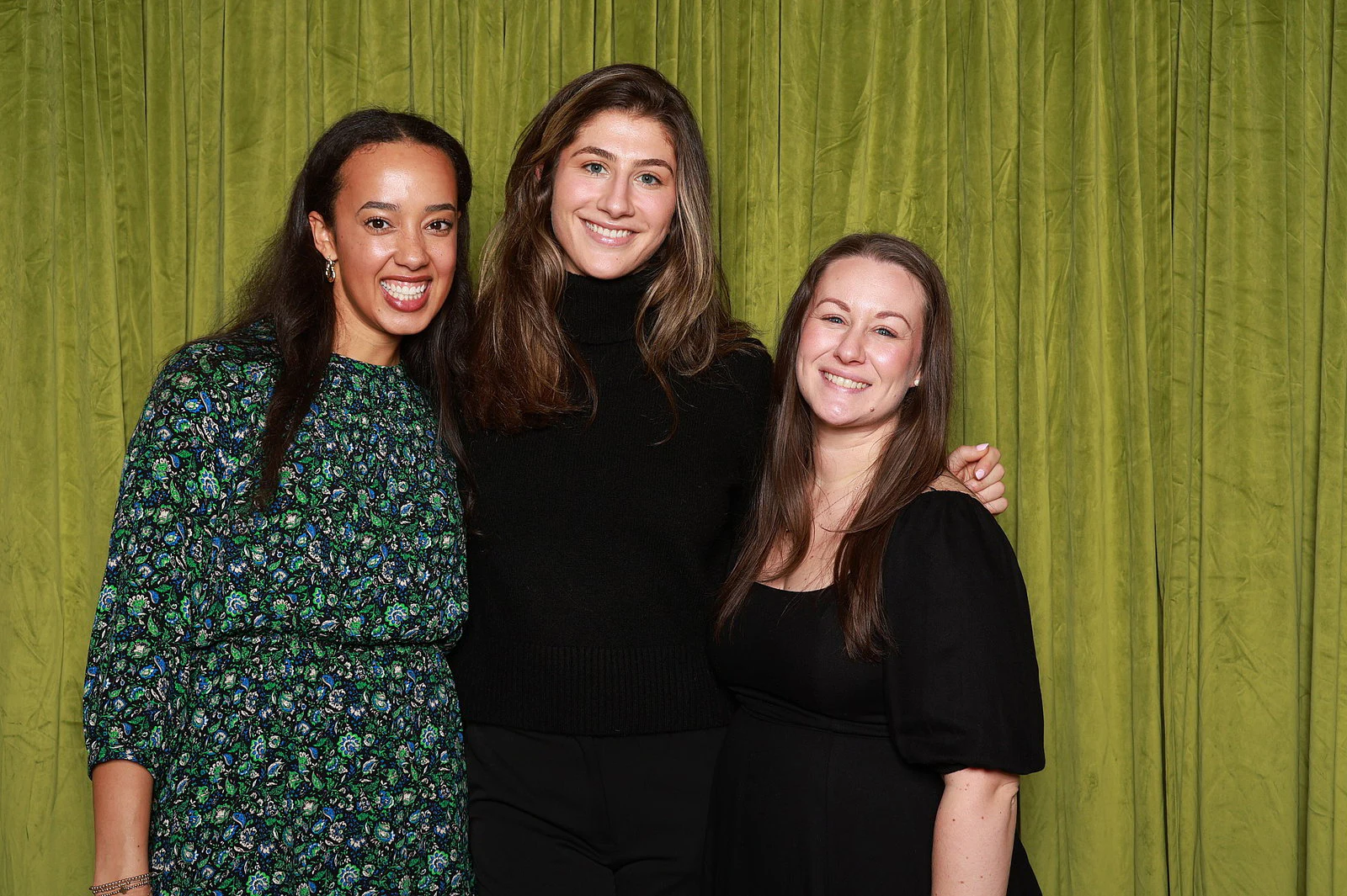 Three women standing close, one in a green print dress and two in black, in front of the olive green velvet at the DYC Tasting