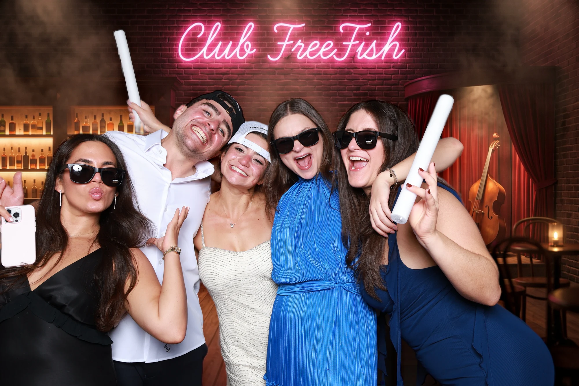 Five guests in sunglasses under the pink neon sign, two waving white light sticks at Eliana + Brad