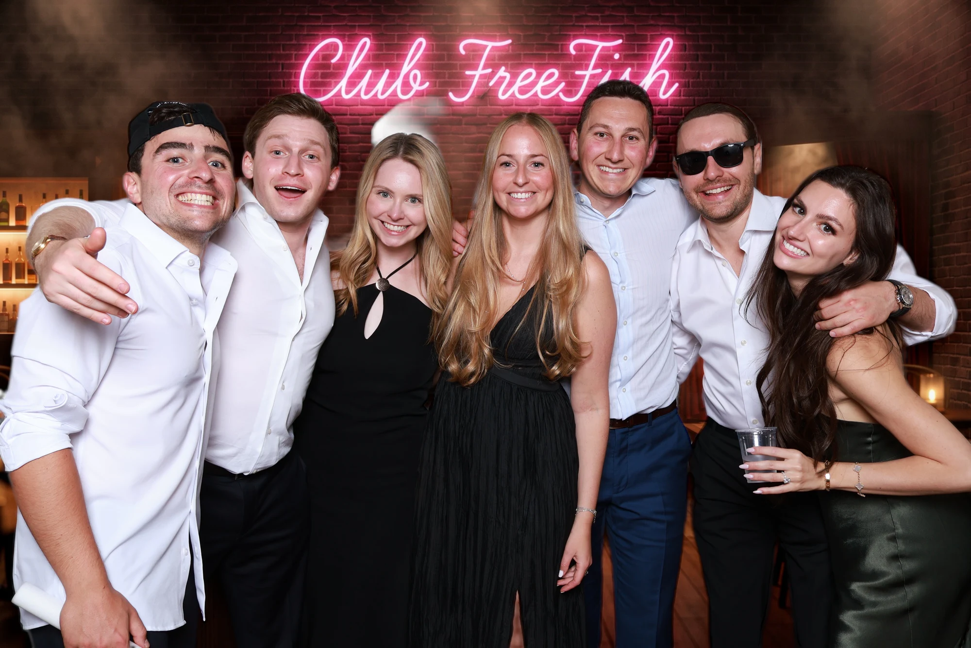 Seven guests in a row under the pink “Club FreeFish” neon sign at Eliana + Brad