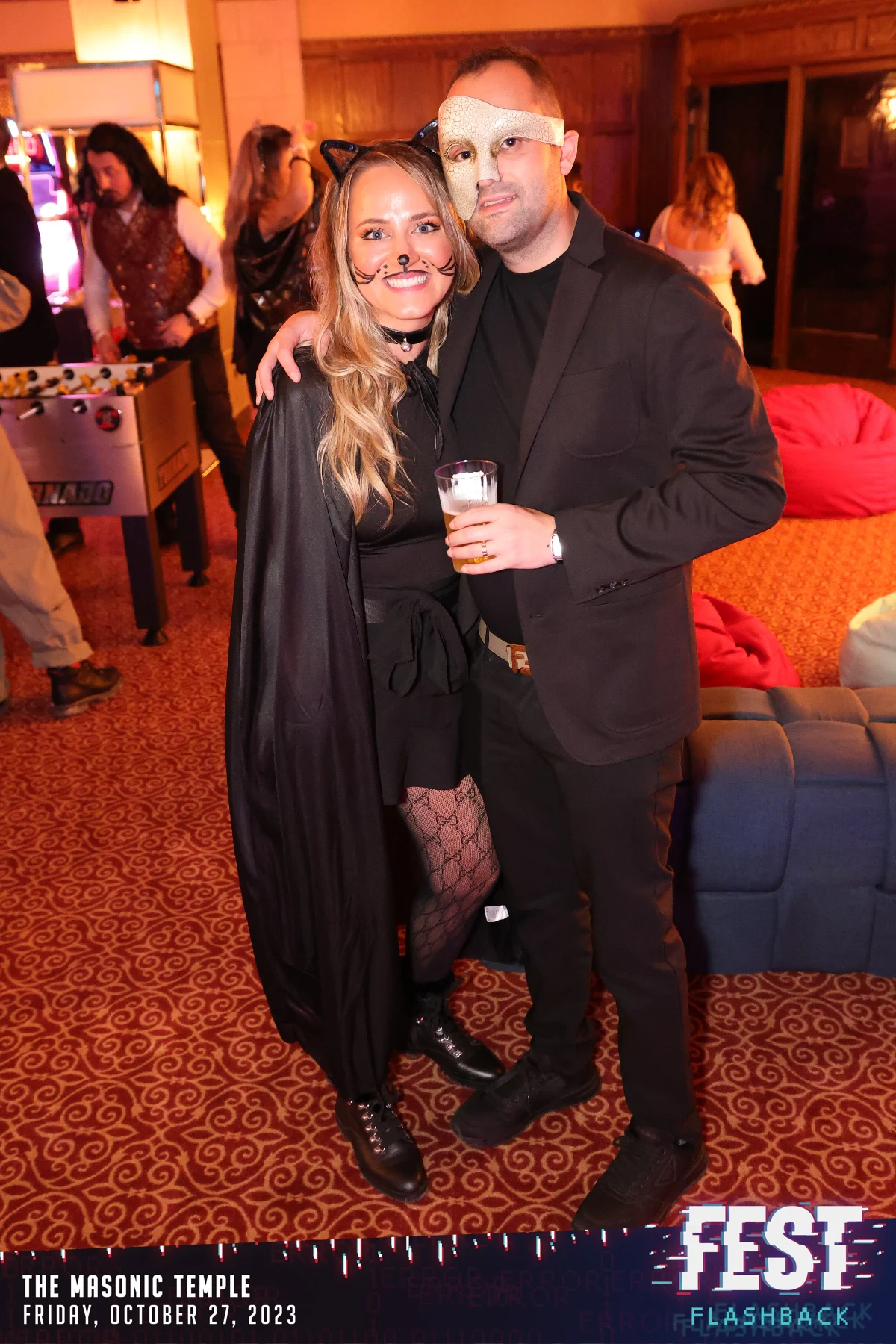 A couple, one with painted cat whiskers and a cape, the other in a masquerade mask at FEST Halloween Party at the Masonic Temple