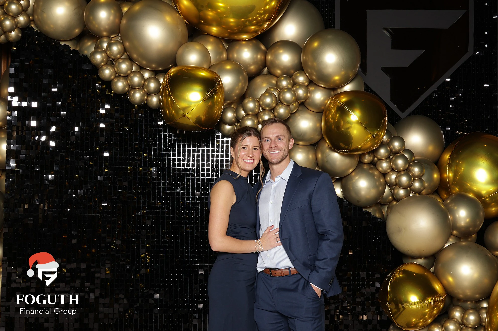 A couple in navy standing close in front of the gold balloons at Foguth Financial Christmas Party