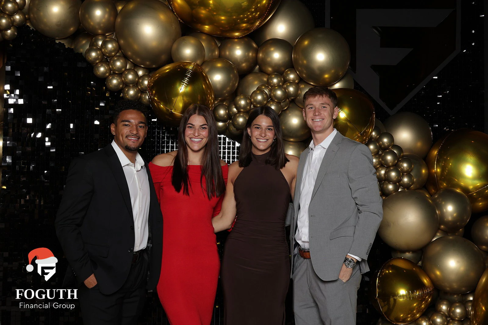 Two men in suits and two women in a red dress and a brown dress in front of a black sequin wall and gold balloons at Foguth Financial Christmas Party
