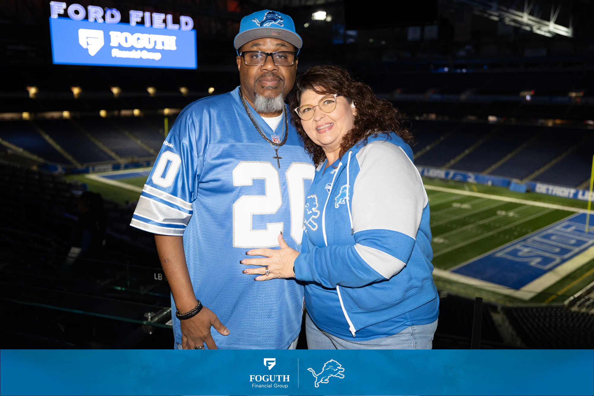 A couple in Lions jerseys with the field behind them at Foguth Financial at Ford Field