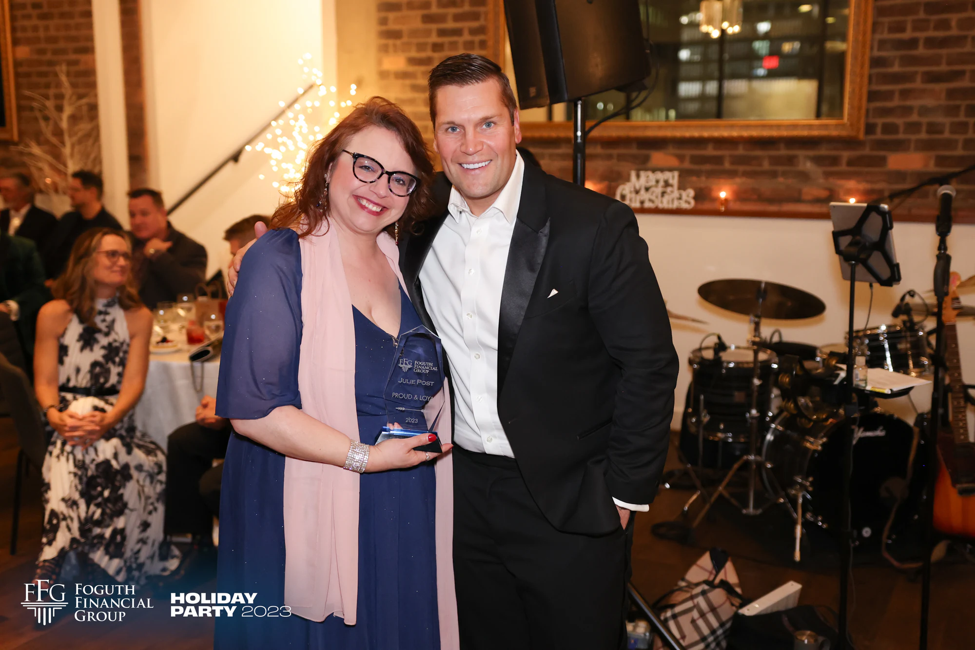 A guest holding a glass award beside a man in a tuxedo, the drum kit behind them at Foguth Financial Holiday Party at the Detroit Foundation Hotel