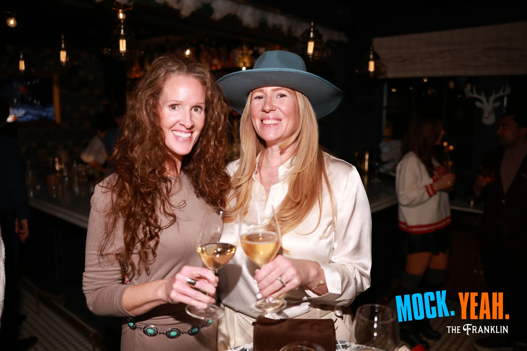 Two guests clinking wine glasses at the bar, one in a pale wide-brim hat at the holiday party at The Franklin