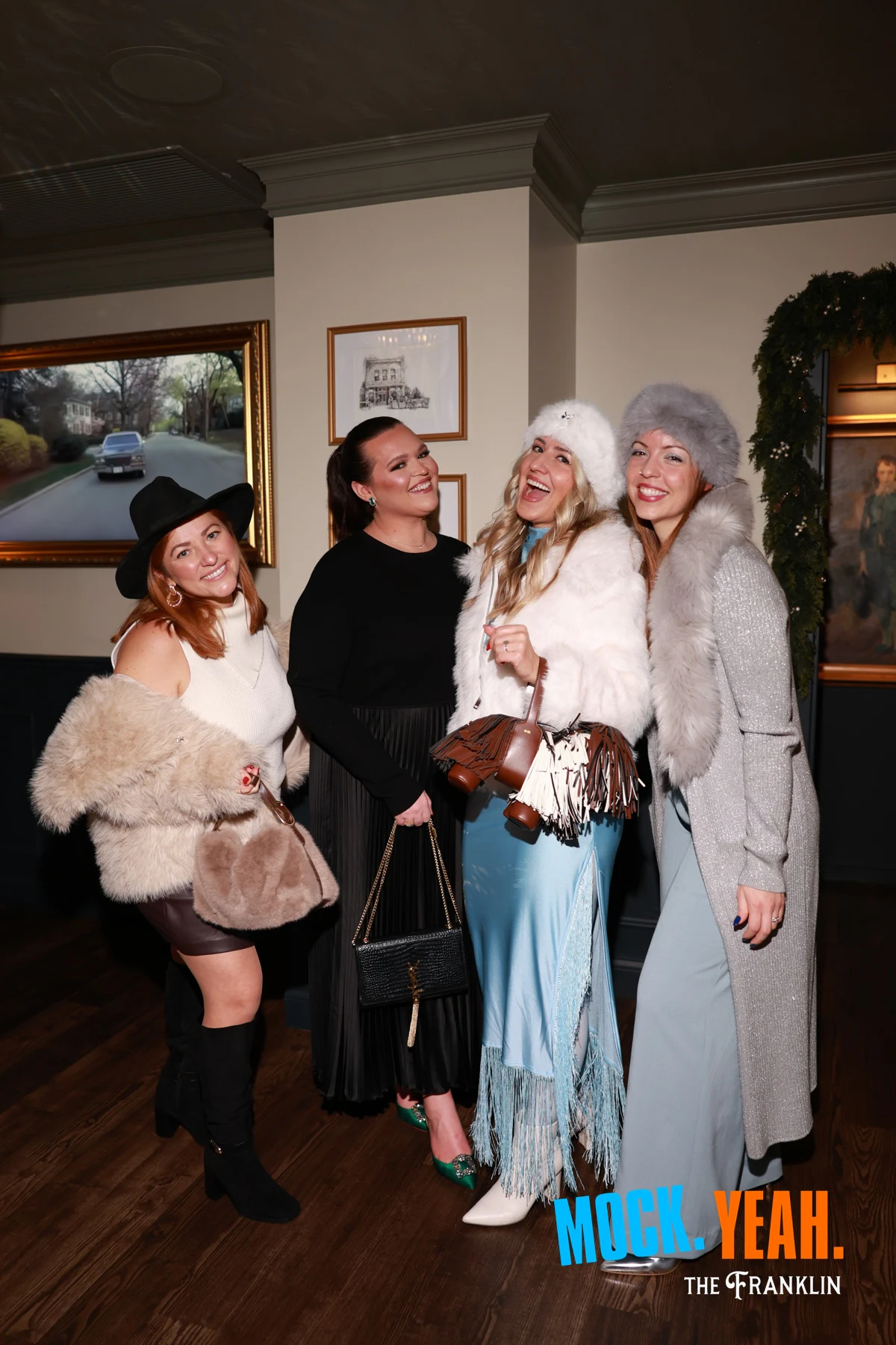 Four guests in fur coats and hats leaning in together at the holiday party at The Franklin