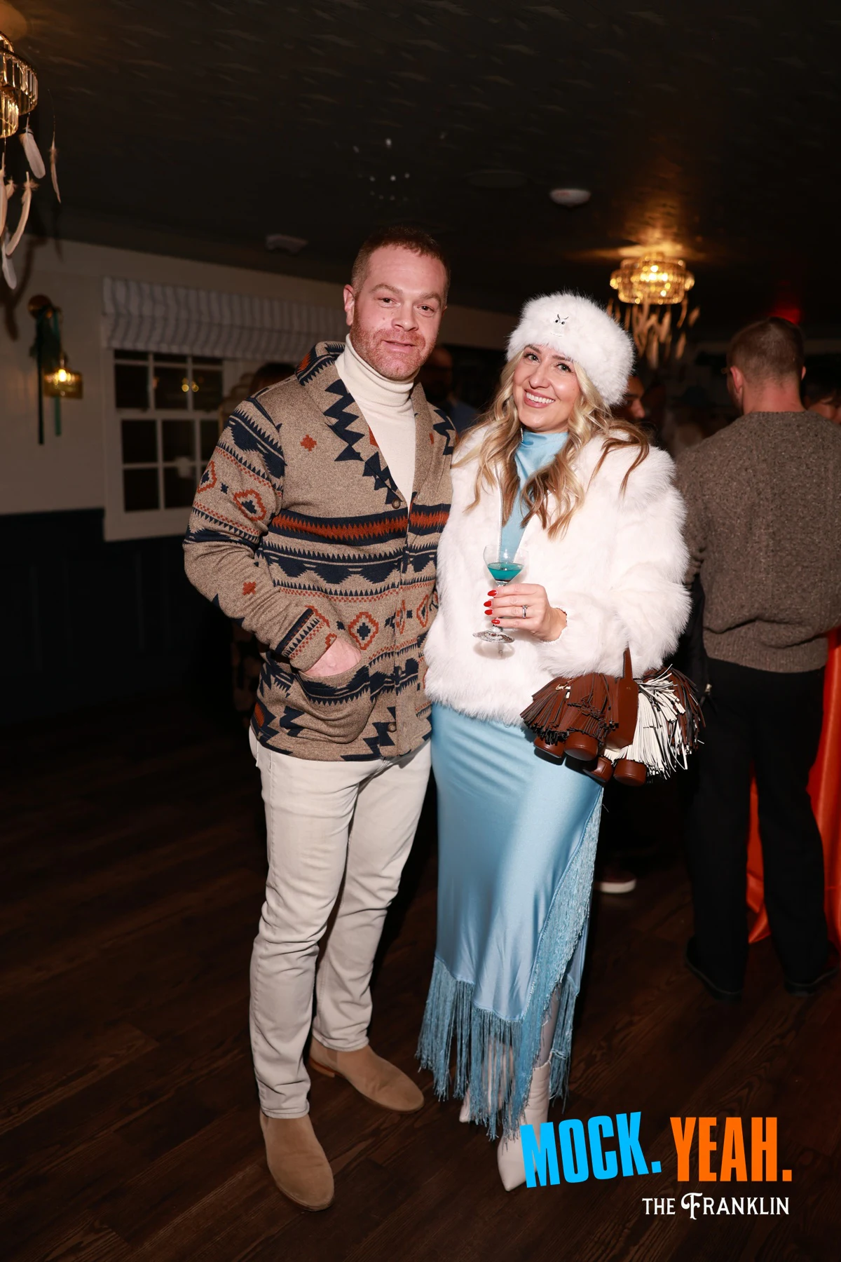 A couple, her in a white fur hat and coat with a cocktail at the holiday party at The Franklin